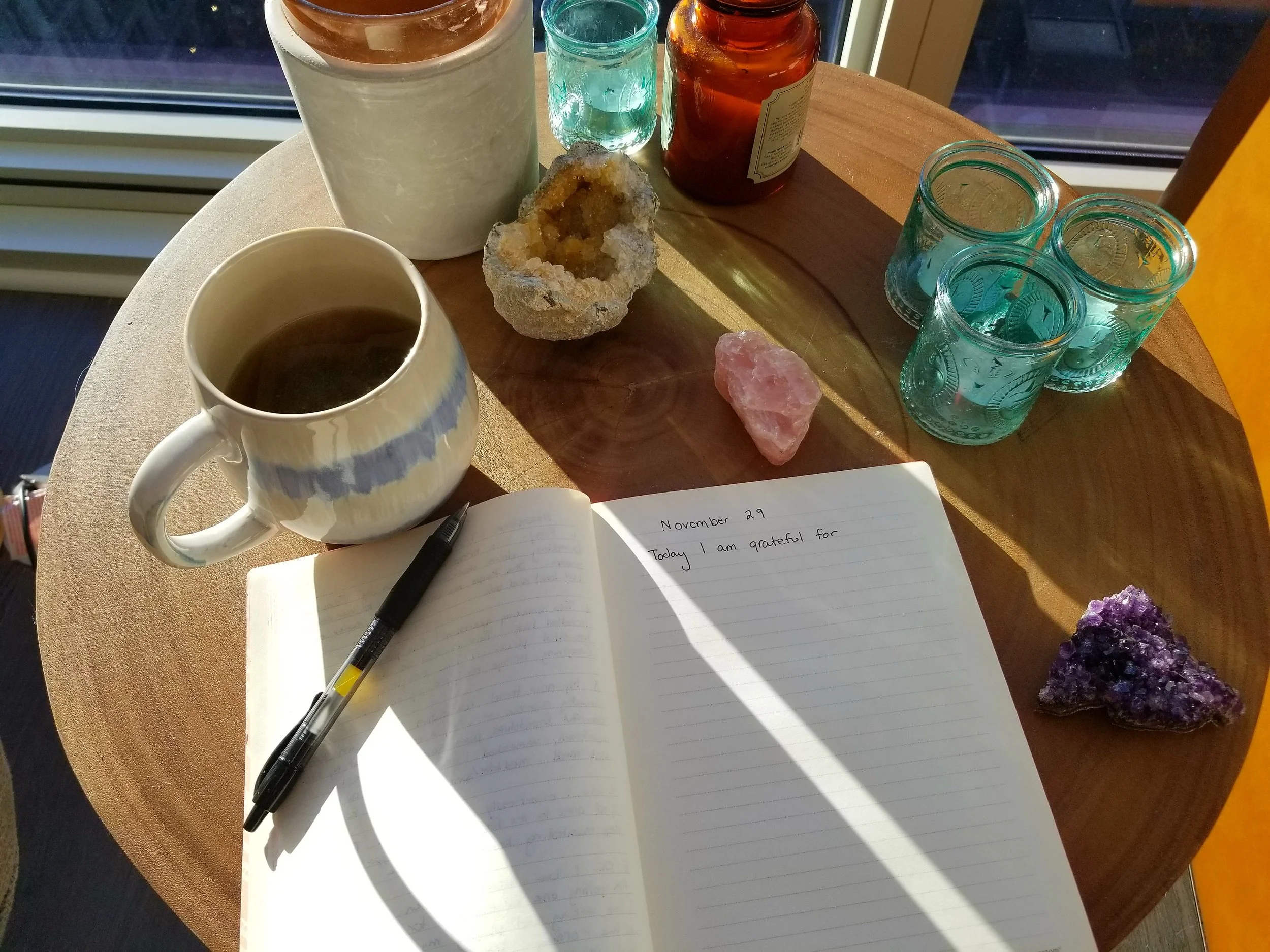 A wooden round table holding a notebook with writing, a black pen, a cup of hot tea, a tall white cup with a beverage, a small teal glass, a glass jar with a brown liquid, a geode, a pink crystal, and a purple mineral. The table is near a window with sunlight streaming in.