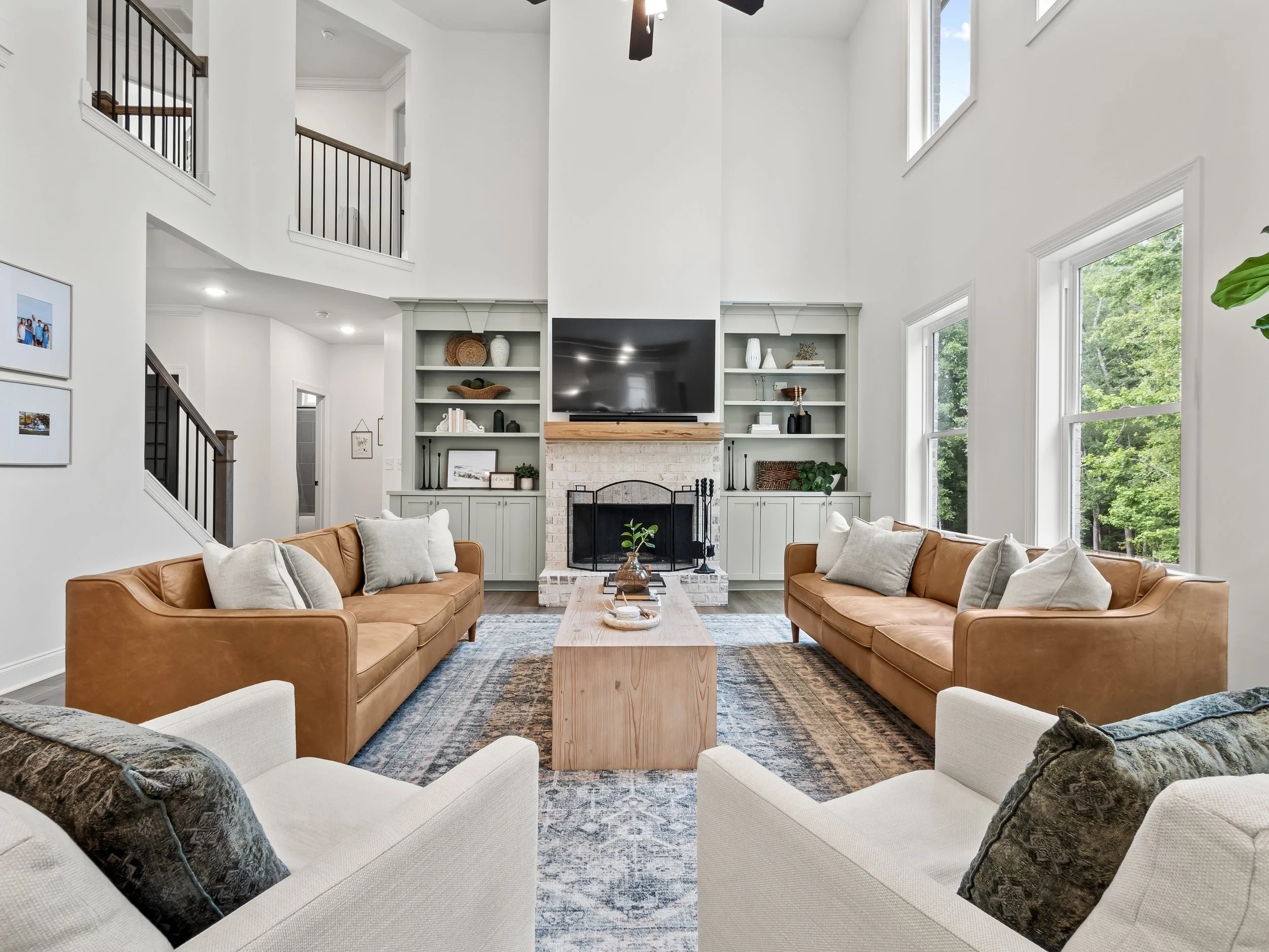 North facing living room with two brown leather sofas, two cream armchairs, a brick fireplace with a wooden mantel, mounted flat-screen TV, built-in shelves, large windows, and a blue patterned rug.