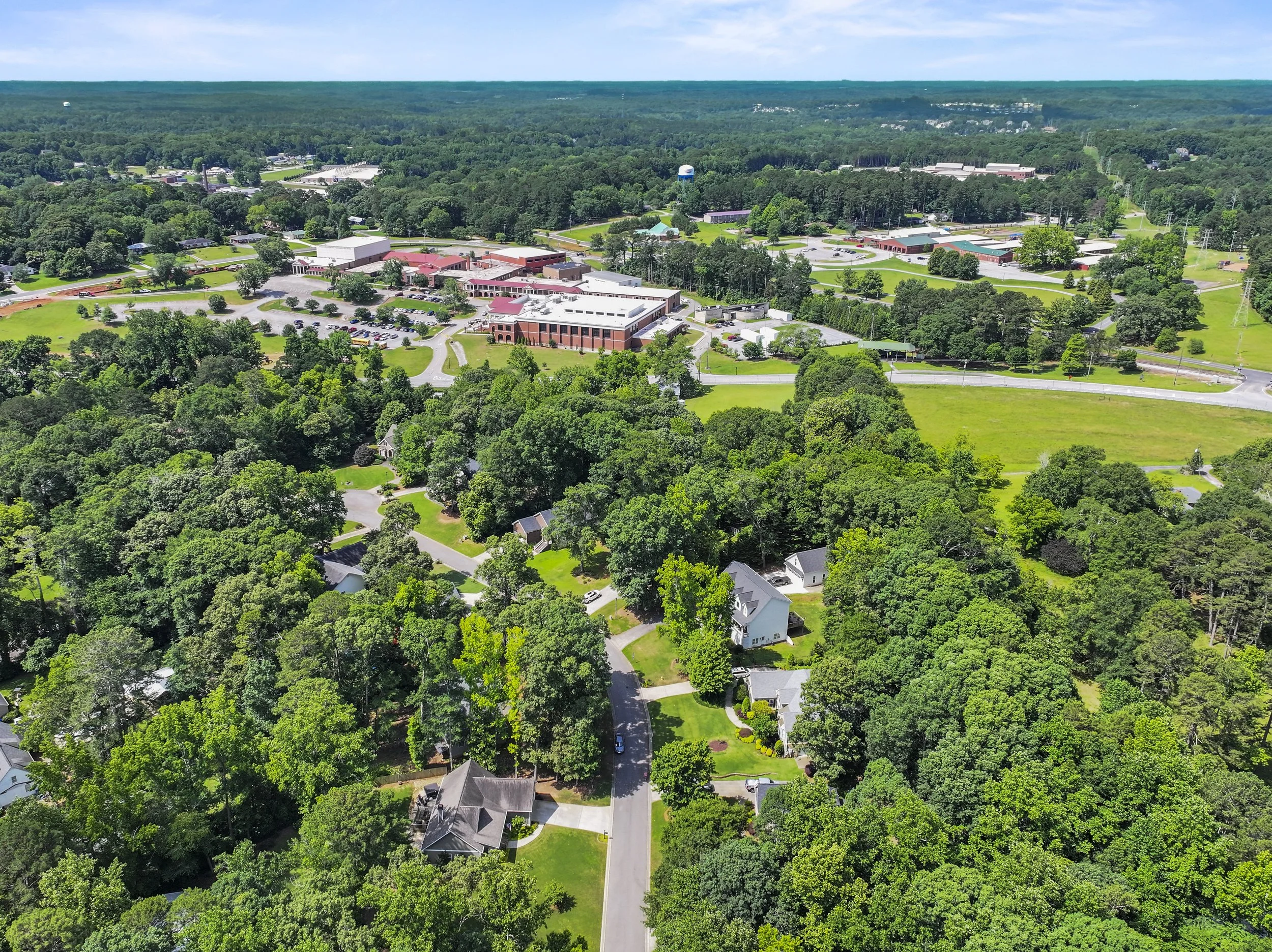 Aerial view of a suburban area with houses, trees, and a large school campus at the edge of a forest, under a partly cloudy sky.