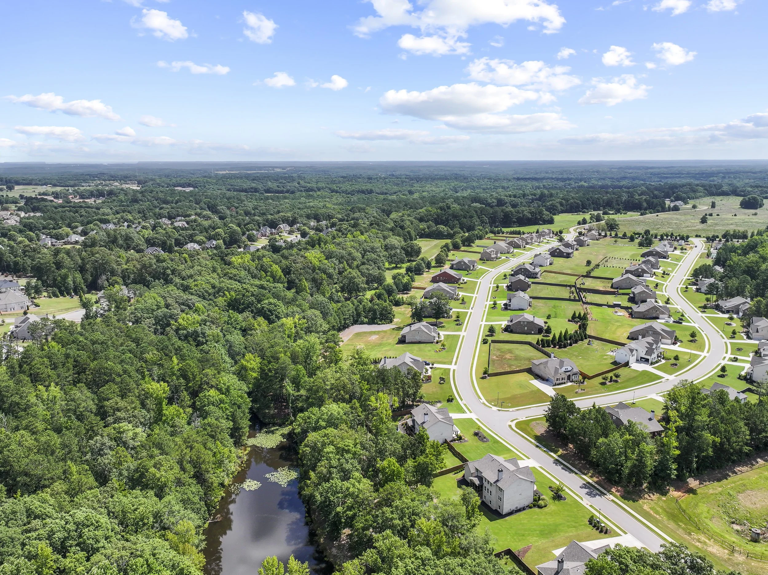 Aerial view of a suburban neighborhood with houses, green lawns, a river, and a large forest under a blue sky with scattered clouds.