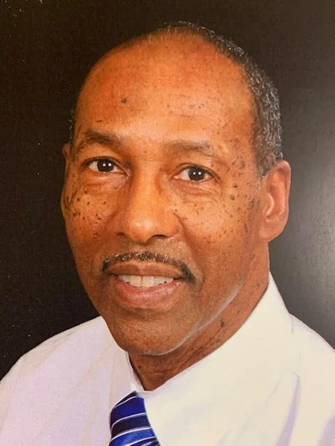 Close-up portrait of a middle-aged African American man with short hair, some facial freckles, wearing a white shirt and a blue striped tie, against a dark background.
