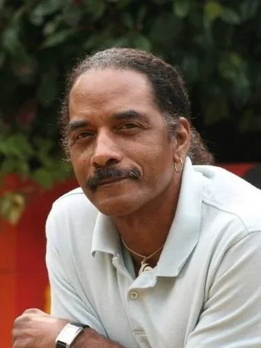 A man with a mustache and dark hair, wearing a light blue polo shirt, sitting outdoors with greenery in the background.