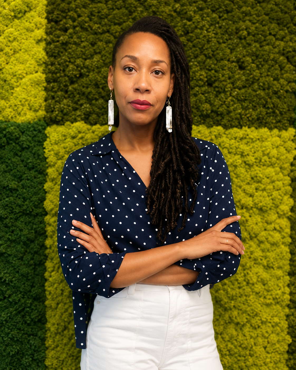 A woman with dark skin, long dreadlocked hair, wearing a navy blue polka dot blouse and white pants, standing with arms crossed in front of a green, leafy textured background.