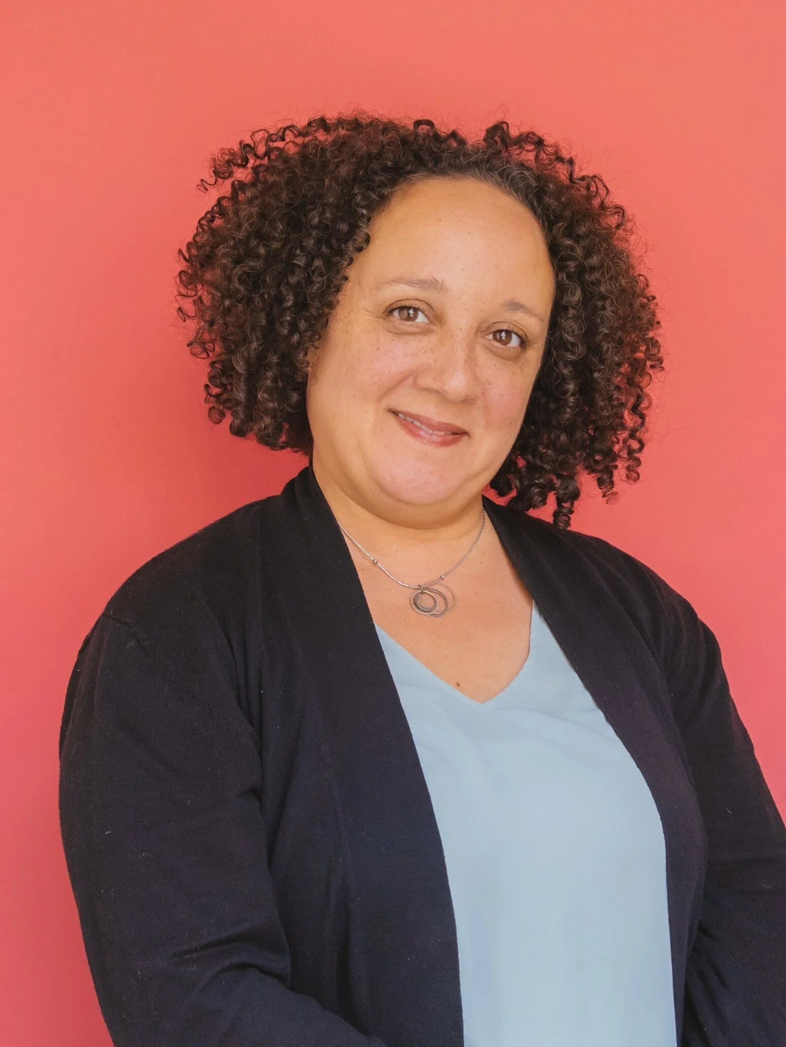 A smiling woman with curly brown hair wearing a light blue top, black cardigan, and a silver necklace, standing in front of a pink background.