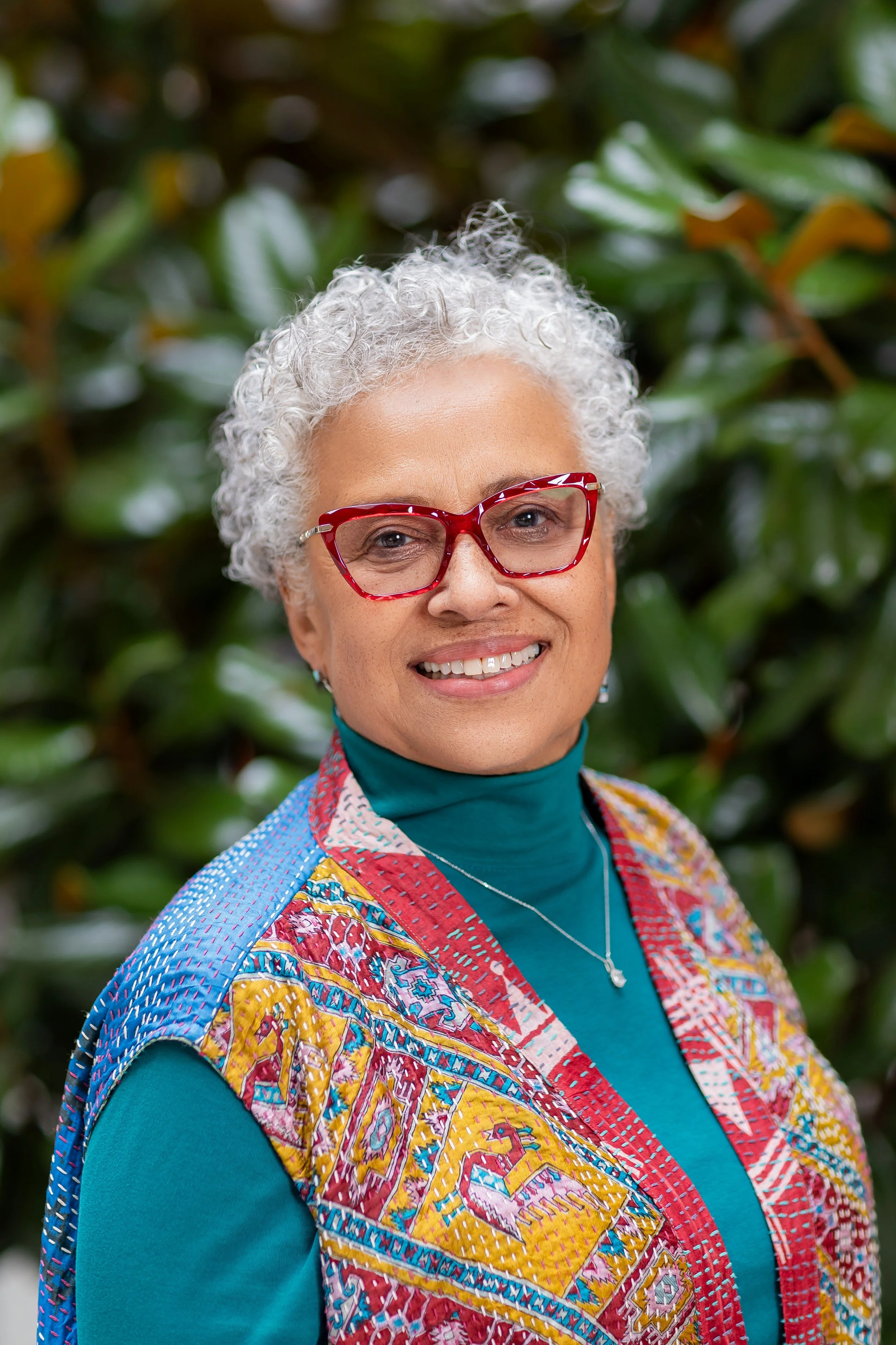 Close-up portrait of a smiling older woman with short, curly gray hair and red glasses, wearing a colorful patterned jacket and a teal turtleneck, standing in front of green leafy background.