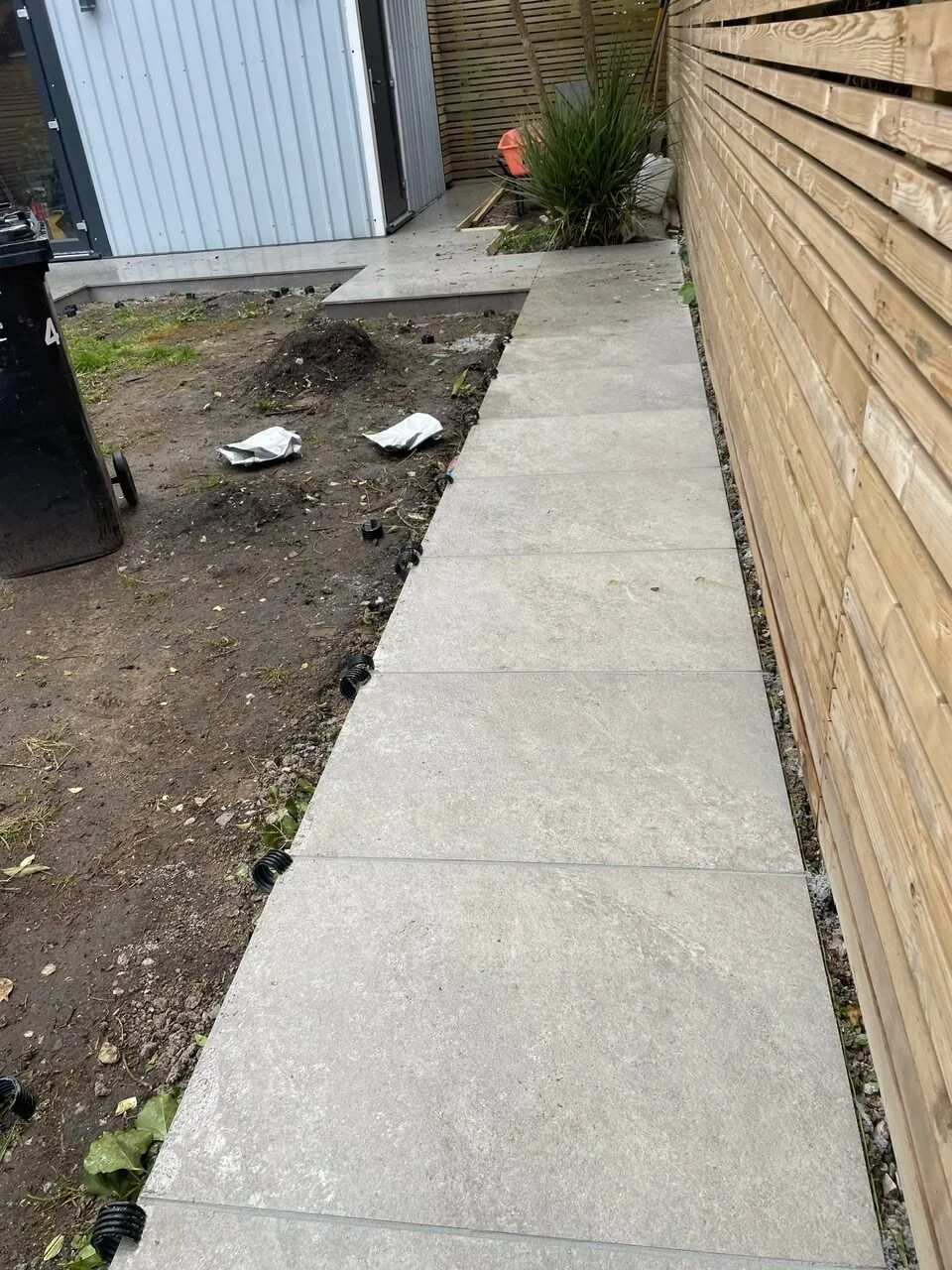 A paved concrete walkway next to a wooden fence, with some soil and construction debris on the side, and a small plant in the background.