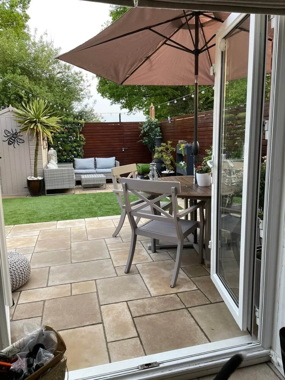 View from inside a house overlooking a backyard patio with a brown umbrella, a round wooden table, and white chairs. The yard has a small artificial grass area, a seating area with a gray outdoor sofa, a shed, potted plants, a tree, and strings of ou
