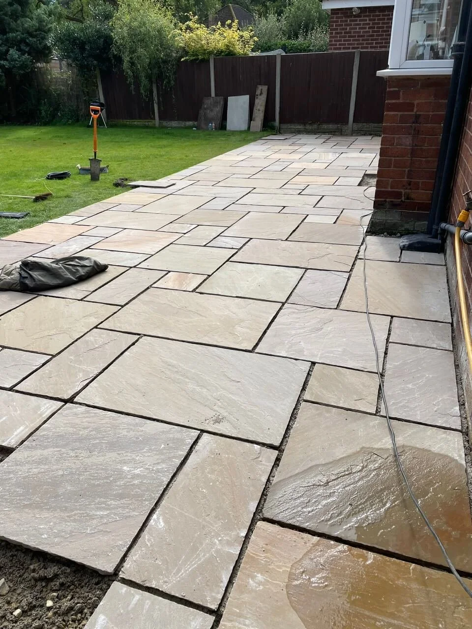 Newly laid stone patio in backyard with some construction tools and materials nearby, next to a grassy lawn and a brick house.