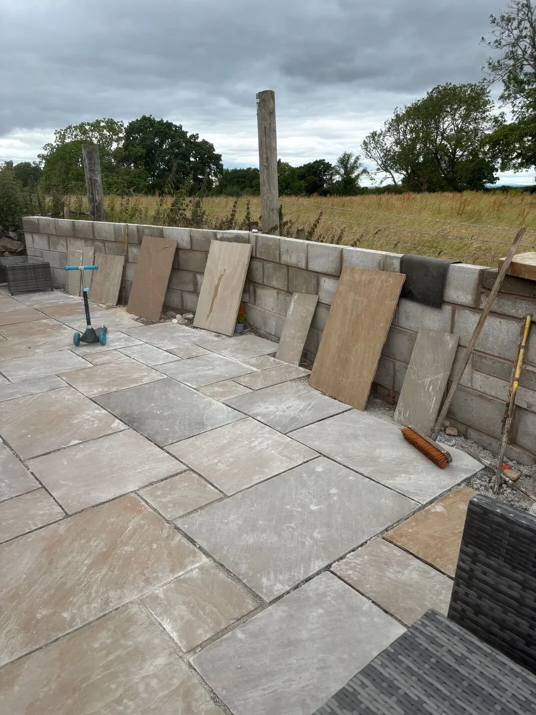 A partially completed outdoor patio with large stone tiles, a stone block wall with some tiles leaning against it, and construction tools like a broom, a scooter, and some stacked bricks. Overcast sky and a field with trees in the background.