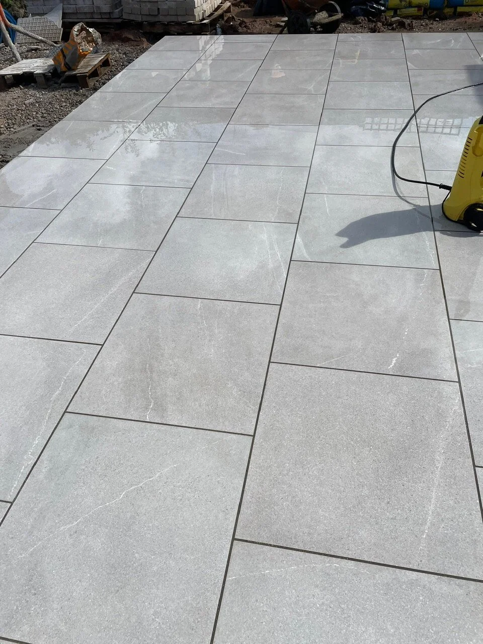 Newly installed gray ceramic tiles on a patio or walkway with a power washer nearby, reflecting sky and surrounding area.