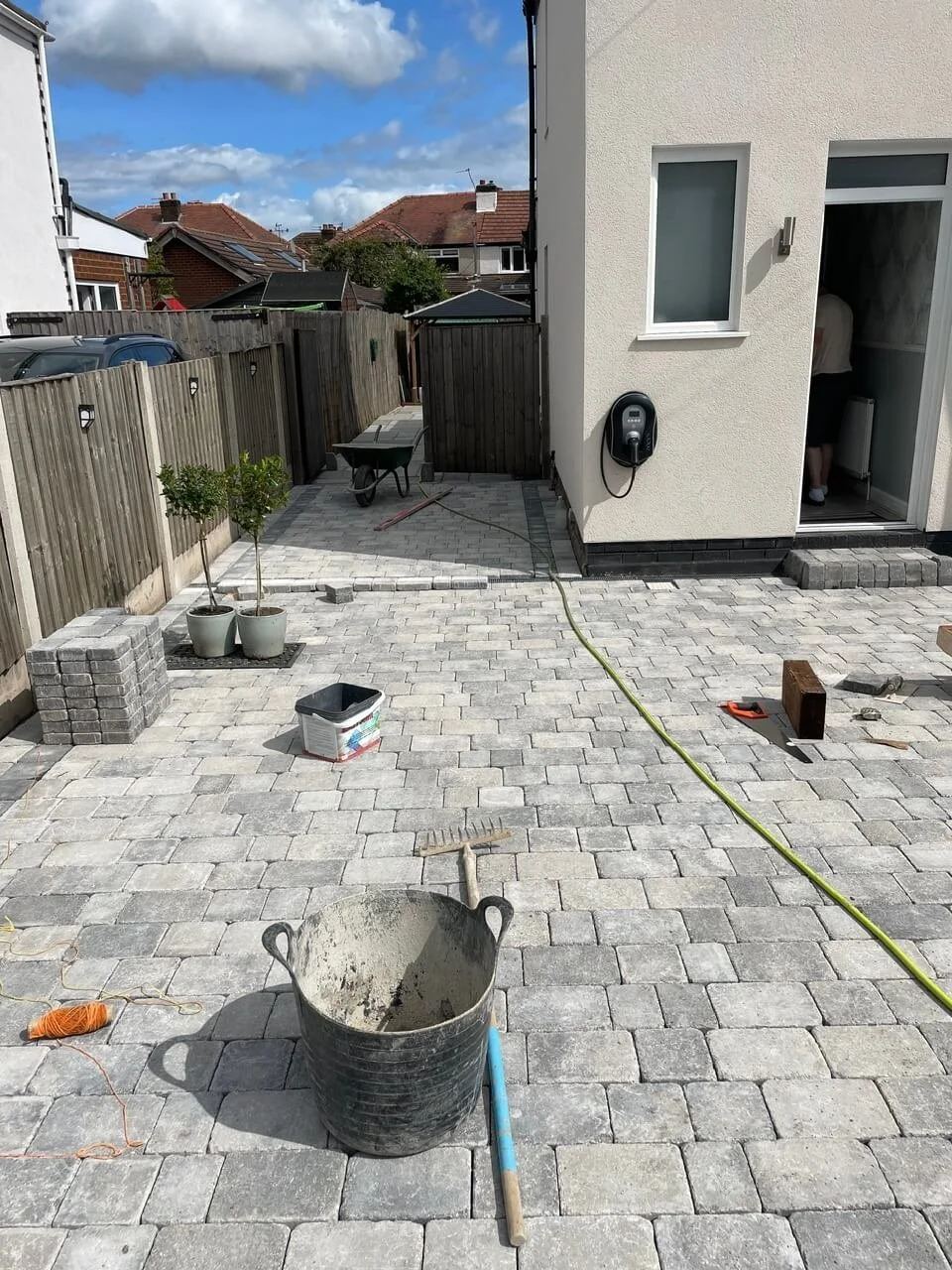 In-progress outdoor patio with paver stones, construction tools, and materials, including a bucket, rake, and plants in pots, adjacent to a white house with an open door and a person inside.