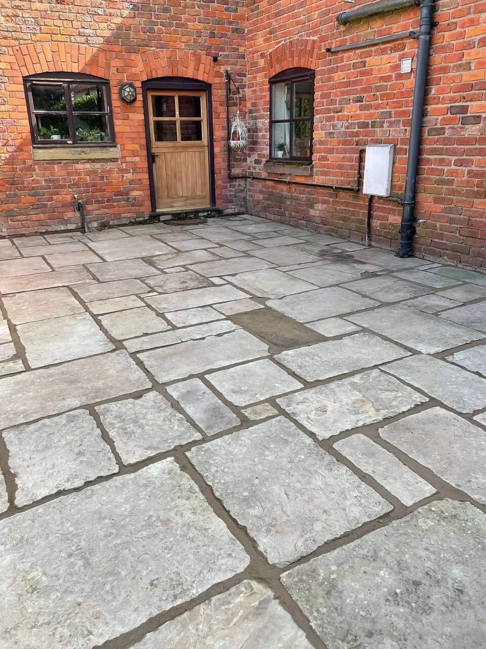 A small outdoor patio area with stone paving in front of a brick house. The house has two windows, a wooden door, and black drains, pipes, and a wall-mounted solar light.