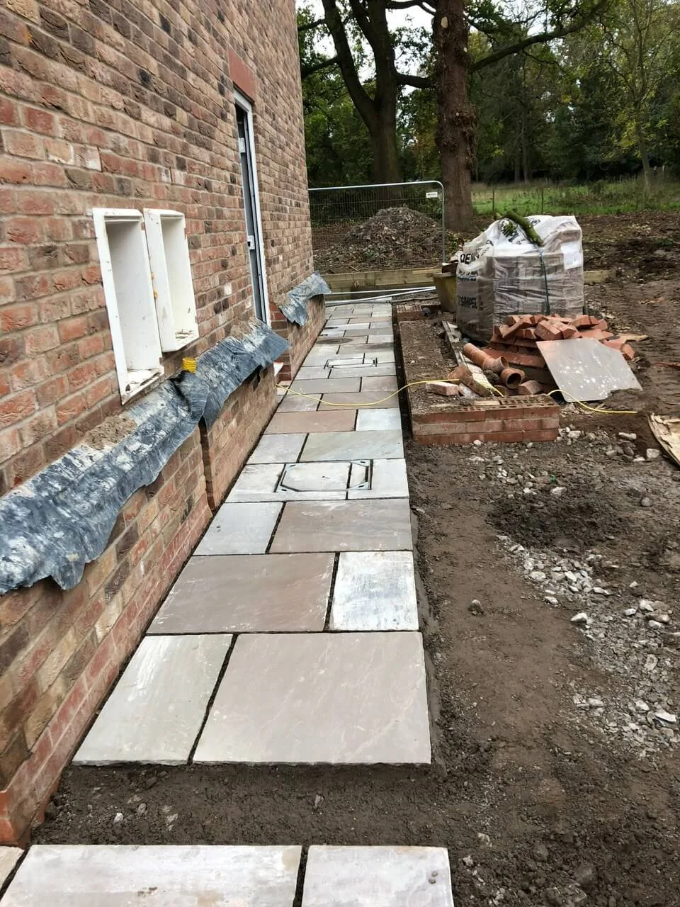 Installation of a stone pathway alongside a brick building with construction materials and tools nearby.