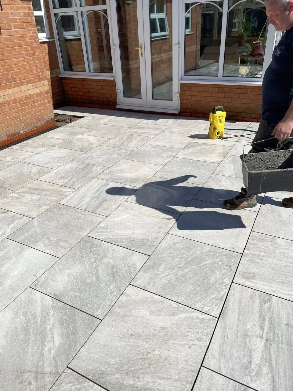 A man standing on a newly installed outdoor tiled patio; the patio has large light gray tiles, and the man is holding a bucket of mortar with a tool. A yellow pressure washer is on the patio, and a glass door with white frames leads into a brick hous