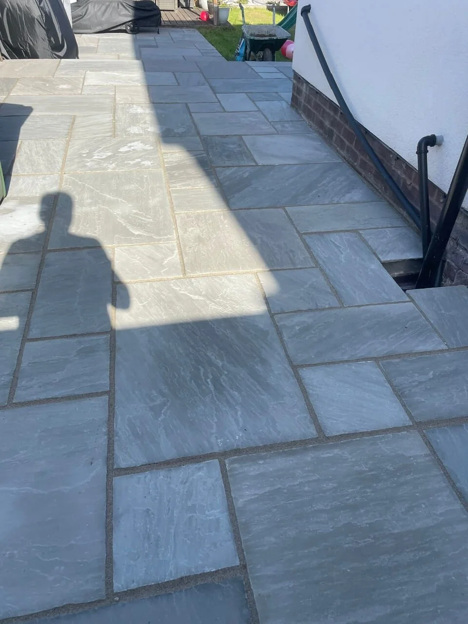 Newly installed gray stone patio with rectangular tiles on a backyard porch, partially in sunlight and shadow, next to a white house with a brick foundation.