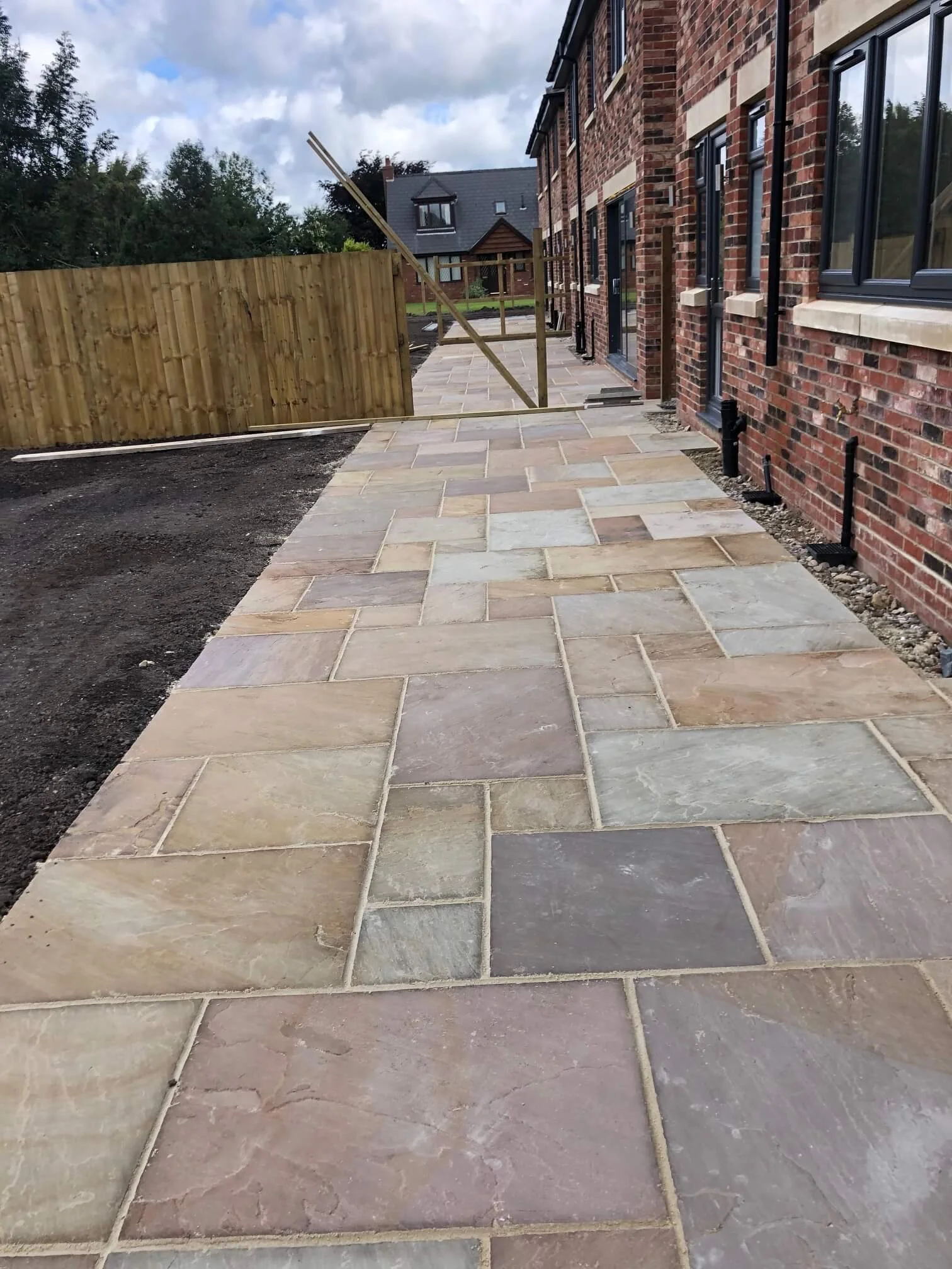 Newly paved stone patio beside a brick house with black windows, with a wooden fence and cloudy sky in the background.