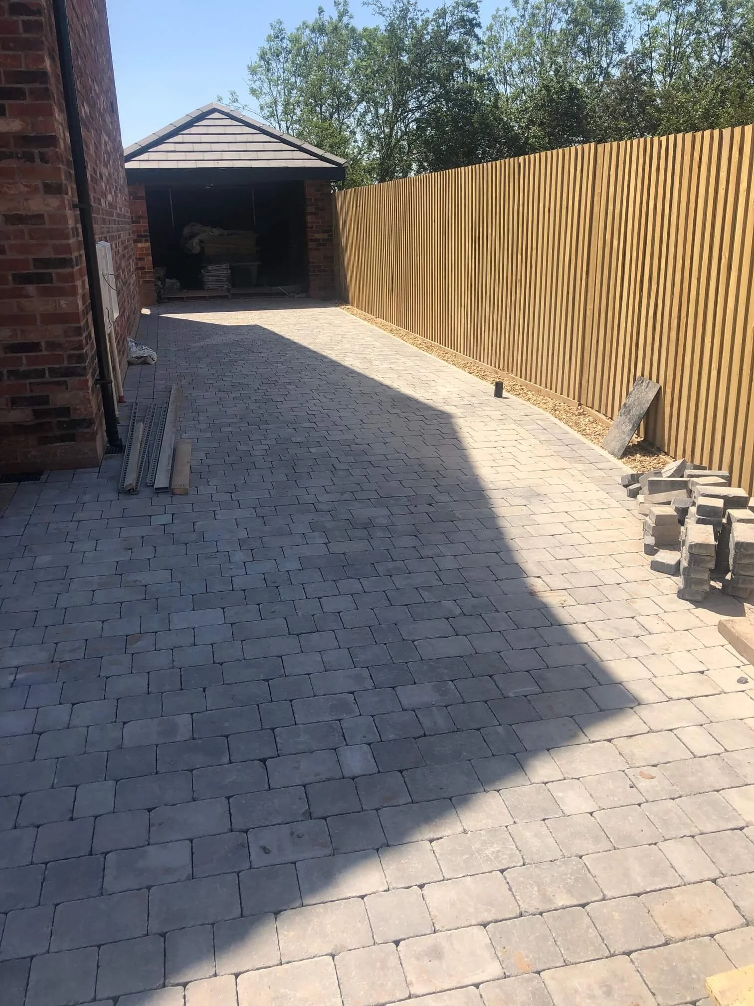 A newly paved driveway with light gray brick pavers, bordered by a wooden fence on the right and a brick house wall on the left, leading to a garage under a small roof with construction materials inside.