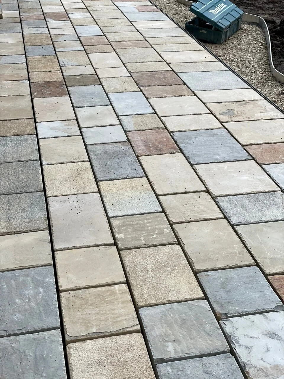 Newly installed stone concrete pavers in various shades of beige, gray, and brown, creating a patterned walking path. Construction materials and tools are visible at the edge of the walkway.