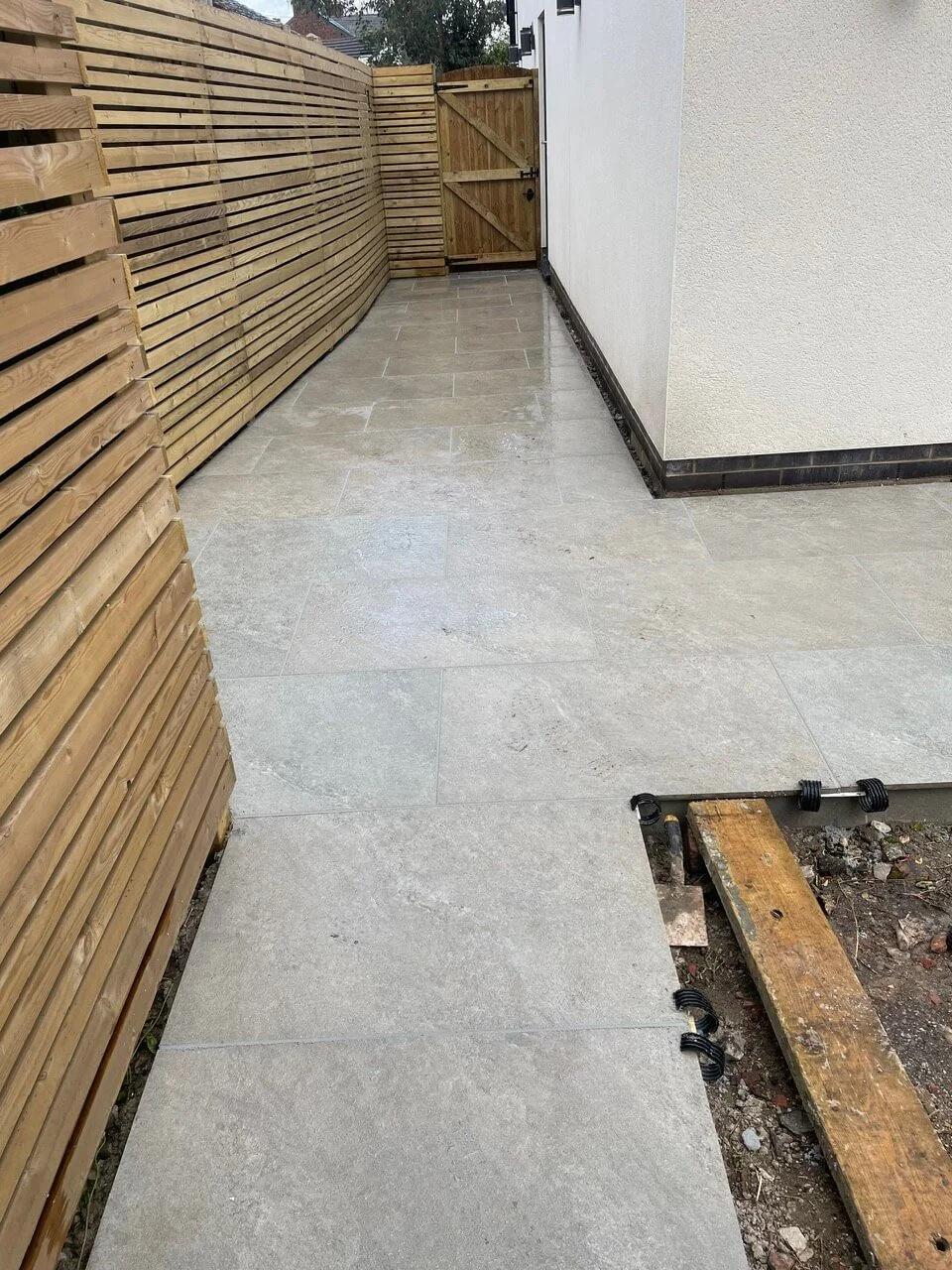 Newly laid outdoor stone tiles on a patio with a wooden fence and a white wall. A wooden plank and some black spacers are visible on the ground, indicating ongoing construction or finishing work.