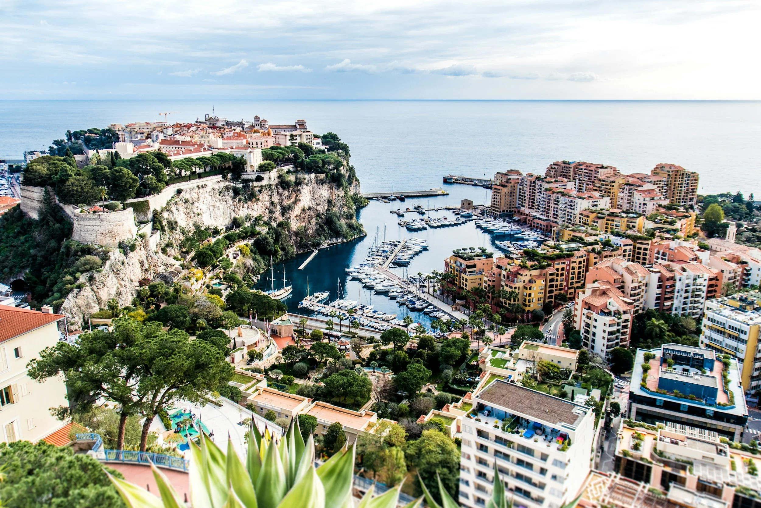 Aerial view of a coastal city with a marina filled with boats, surrounded by colorful residential buildings and a historic fortress on a hill, overlooking the sea.