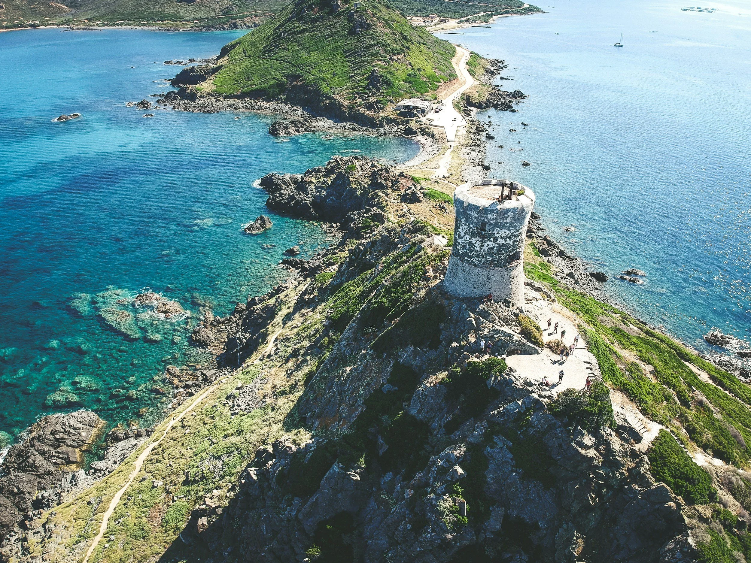 A scenic aerial view of a coastal landscape featuring a historic stone tower on a rocky hill, surrounded by lush green vegetation and overlooking the turquoise ocean, with a small boat in the distance