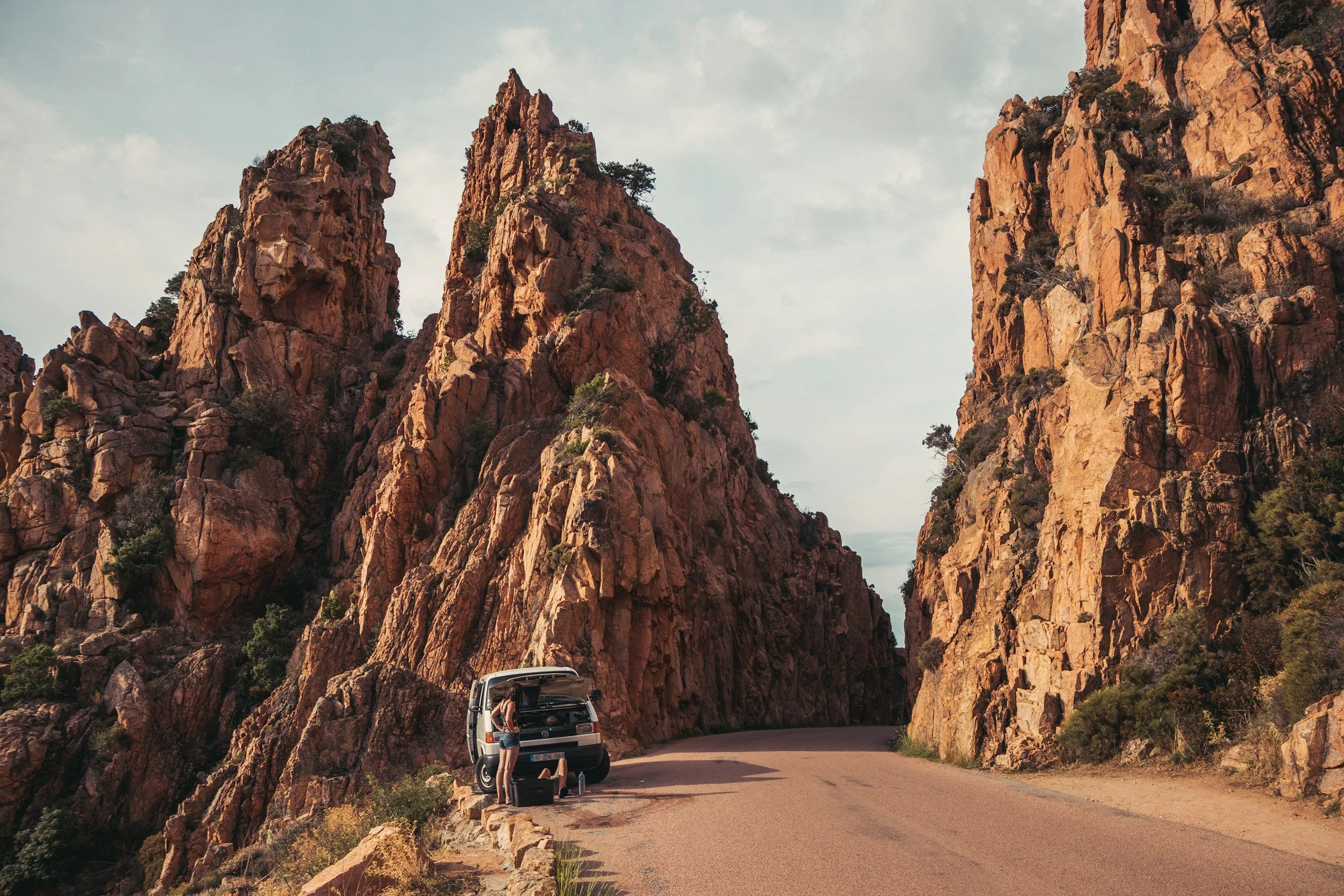 A person standing next to a vehicle with the trunk open, on a mountain road carved through tall, rugged orange rock formations with sparse greenery.