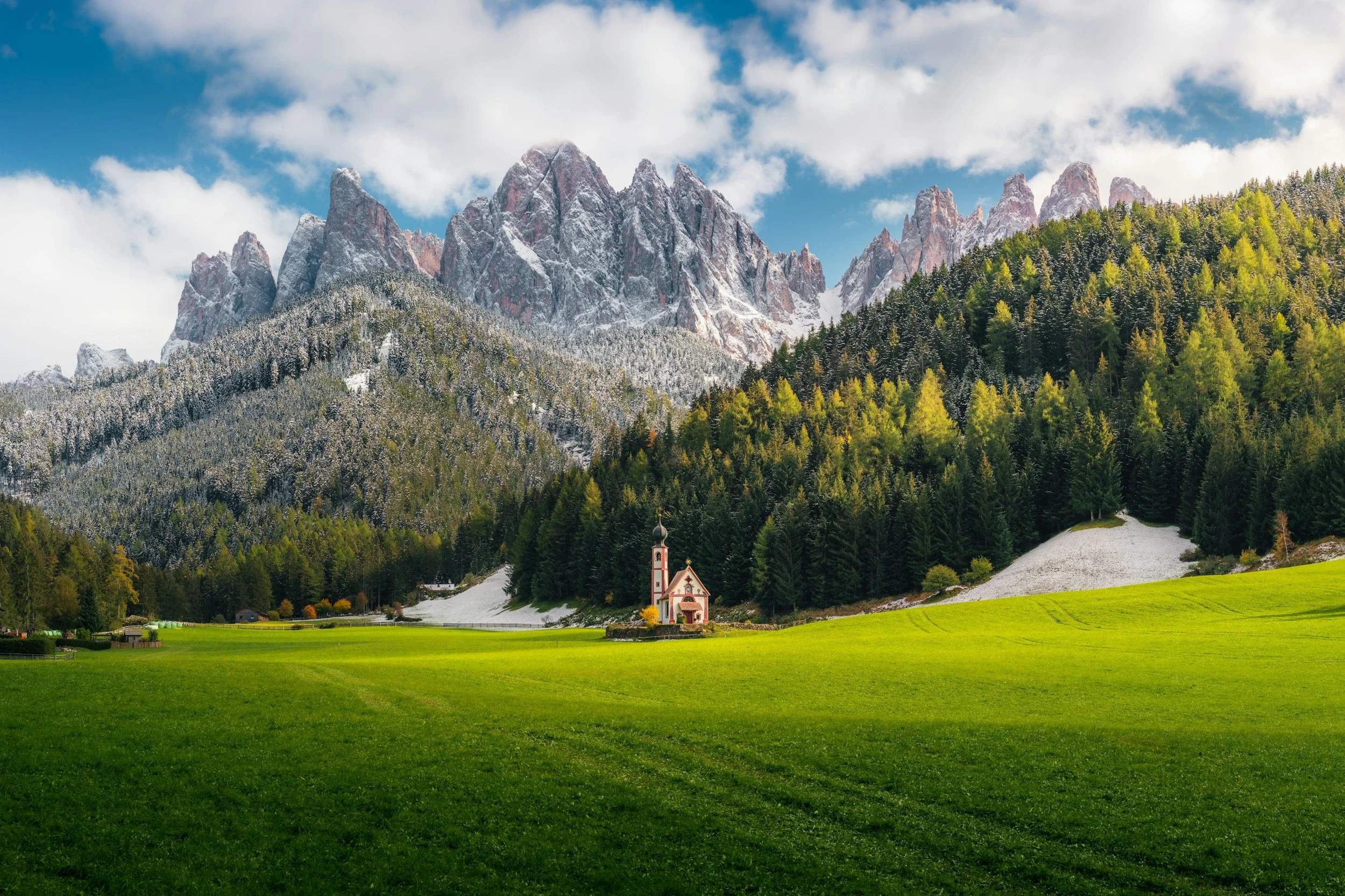 A lush green field with a small church in front of a forested mountain range, some snow on the peaks, under a partly cloudy sky.