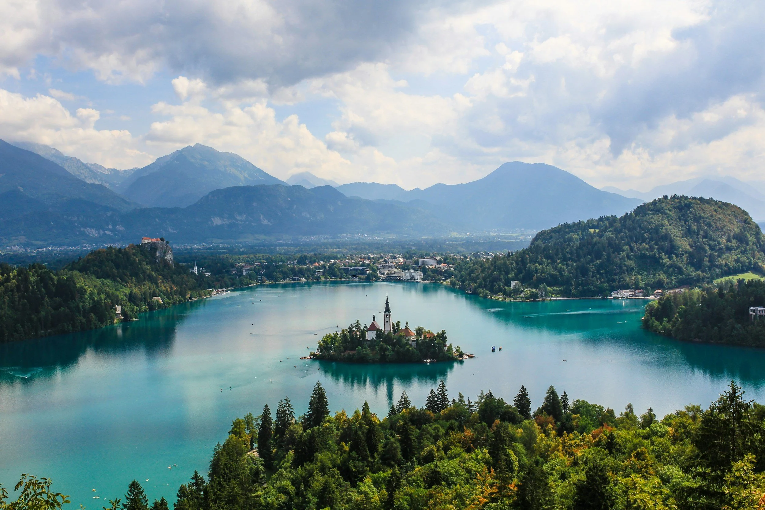 A scenic view of a lake surrounded by lush green hills and mountains in the background, with a small island in the center featuring a church with a tall steeple.