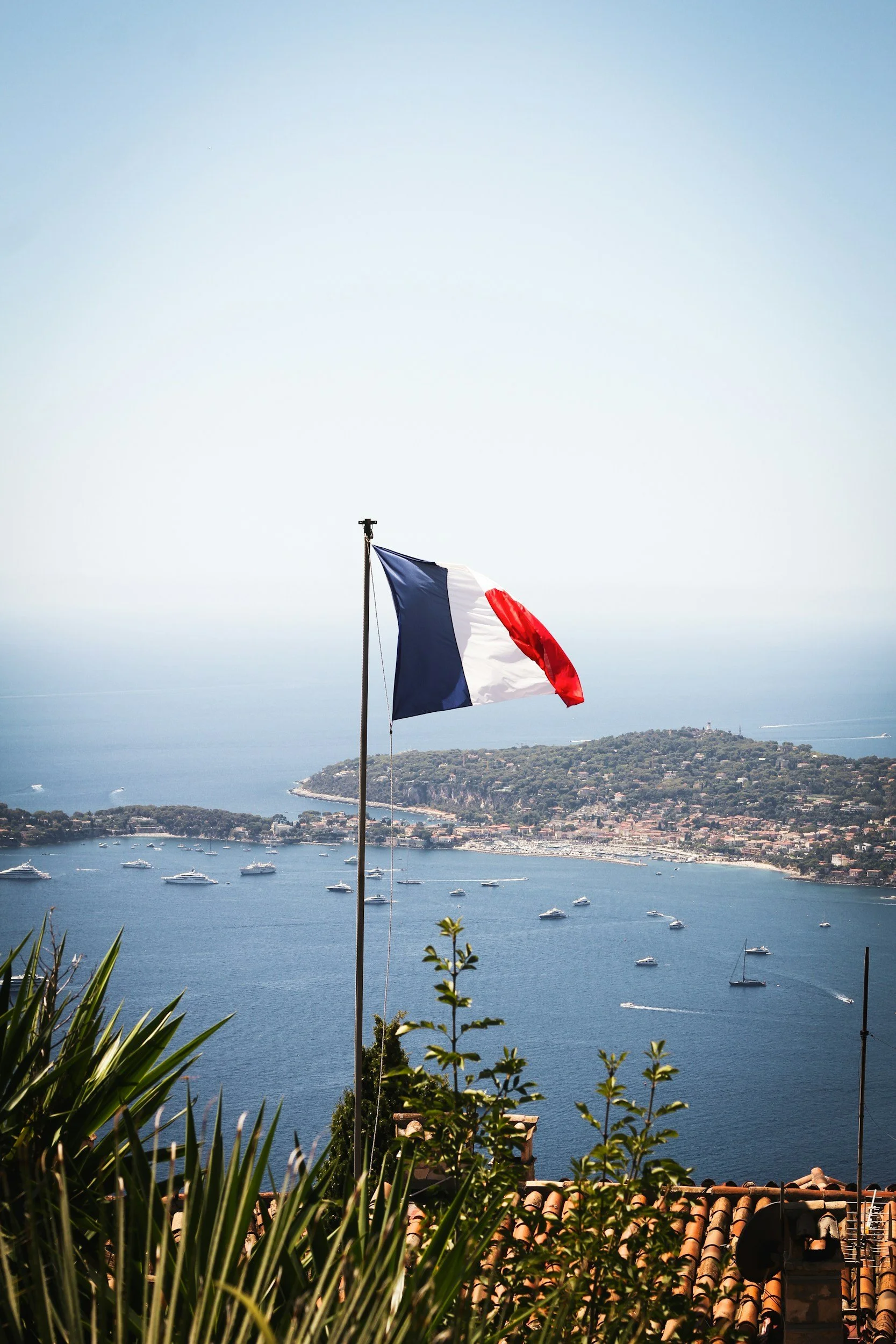 French flag flying above rooftops overlooking a harbor with numerous boats and an island in the distance