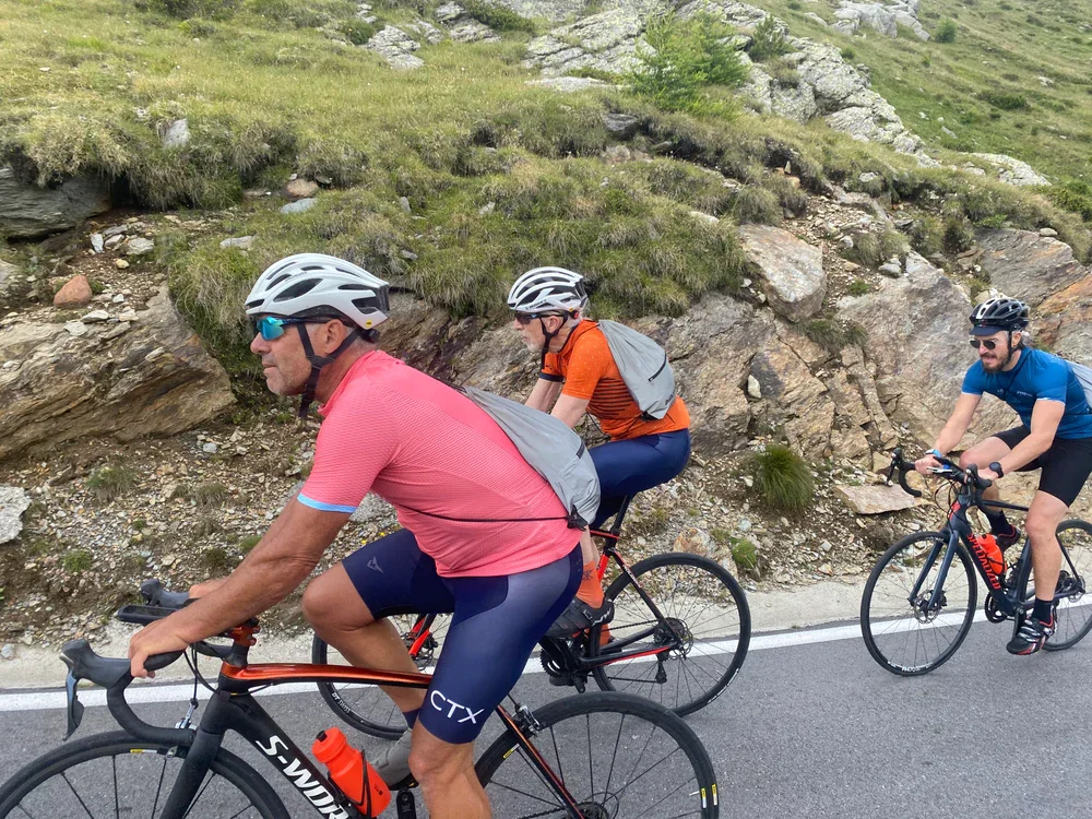 Three cyclists riding on a mountain road with rocky terrain and green vegetation in the background.