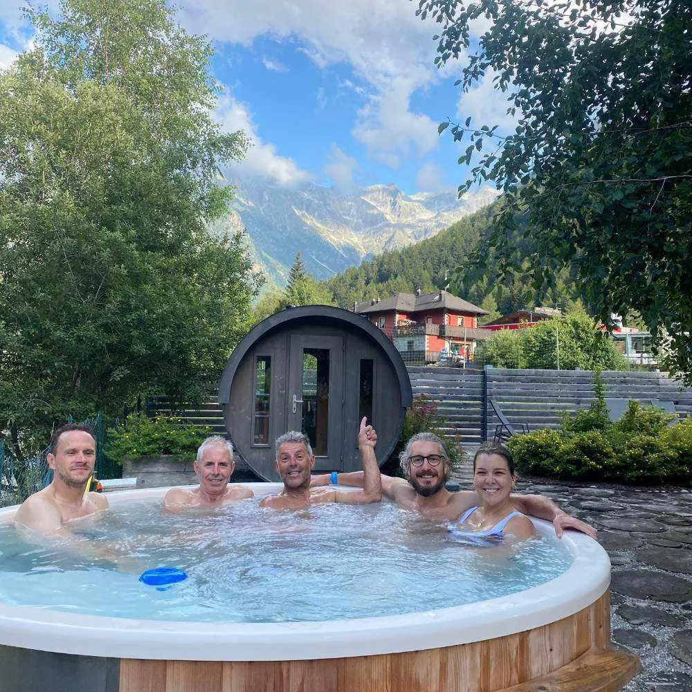 Five people relaxing in an outdoor hot tub with mountains and trees in the background.