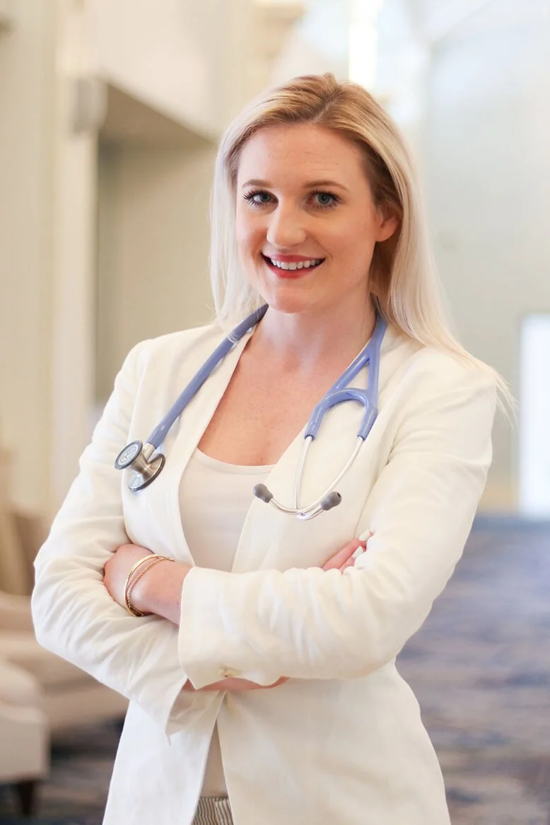 A female doctor with long blonde hair, wearing a white coat and stethoscope, smiling with arms crossed in a medical facility.