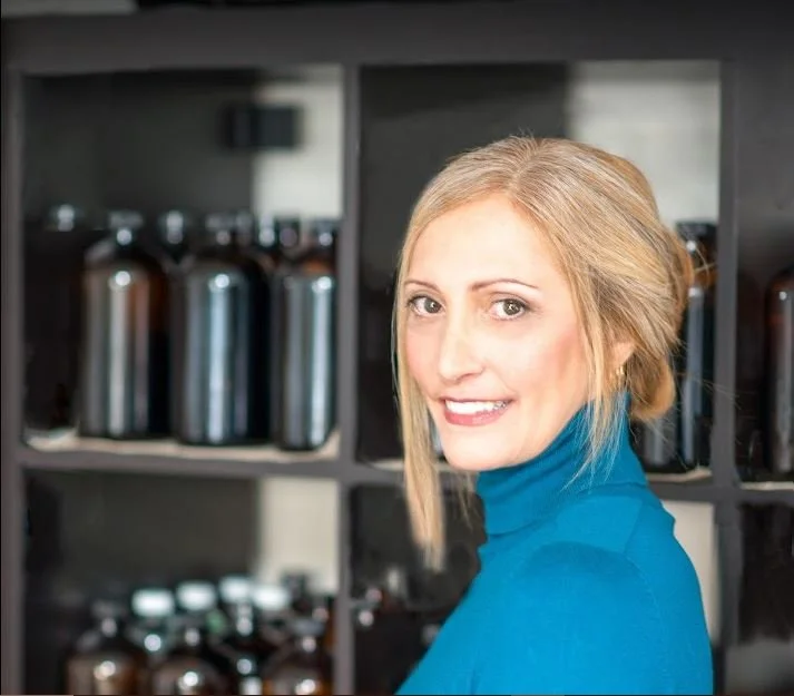 A woman with blonde hair wearing a blue turtleneck, smiling, standing in front of a shelf with black bottles.
