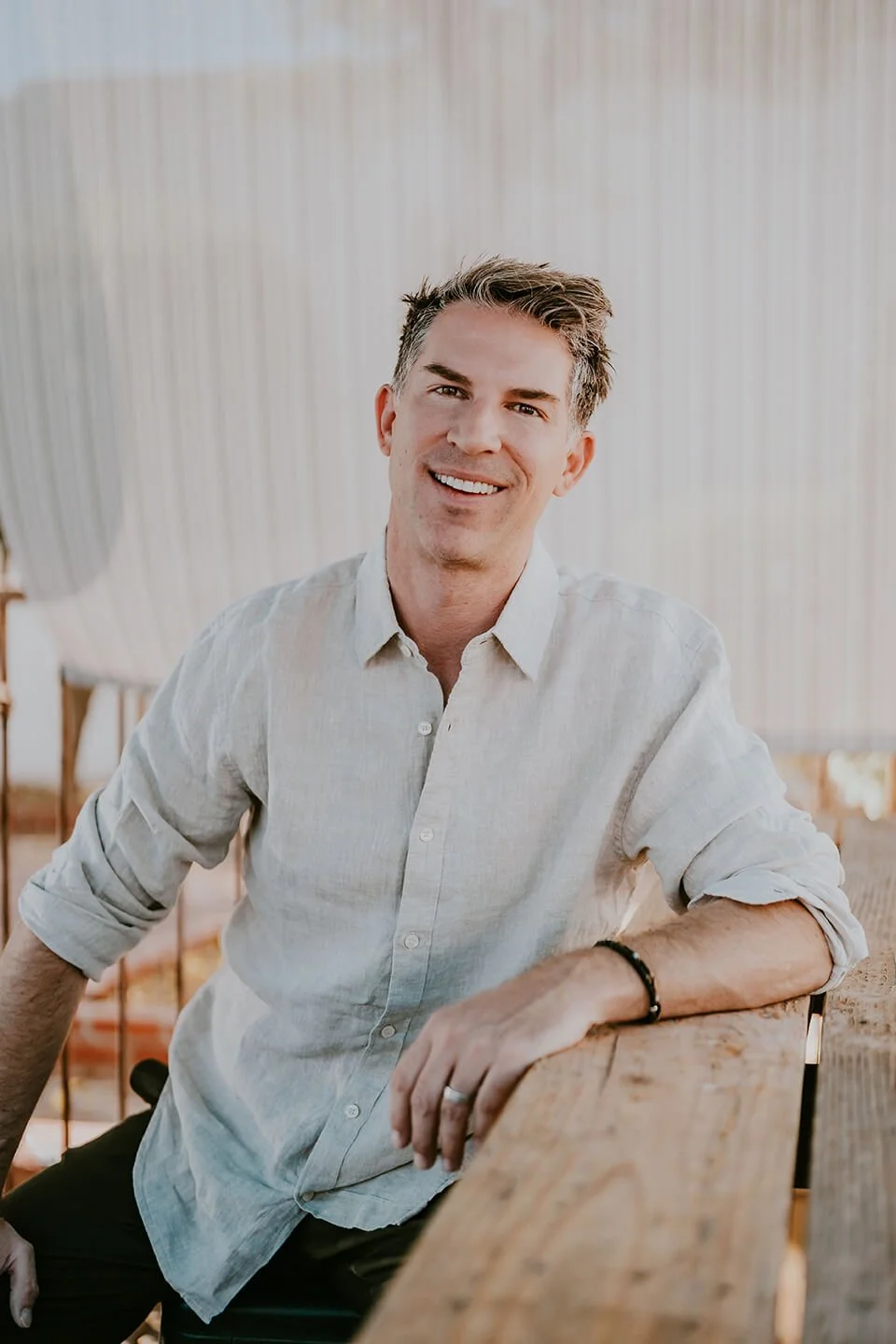 A smiling man with short, styled brown hair, wearing a light gray button-up shirt with the sleeves rolled up, sitting casually at a wooden table in a bright indoor setting.