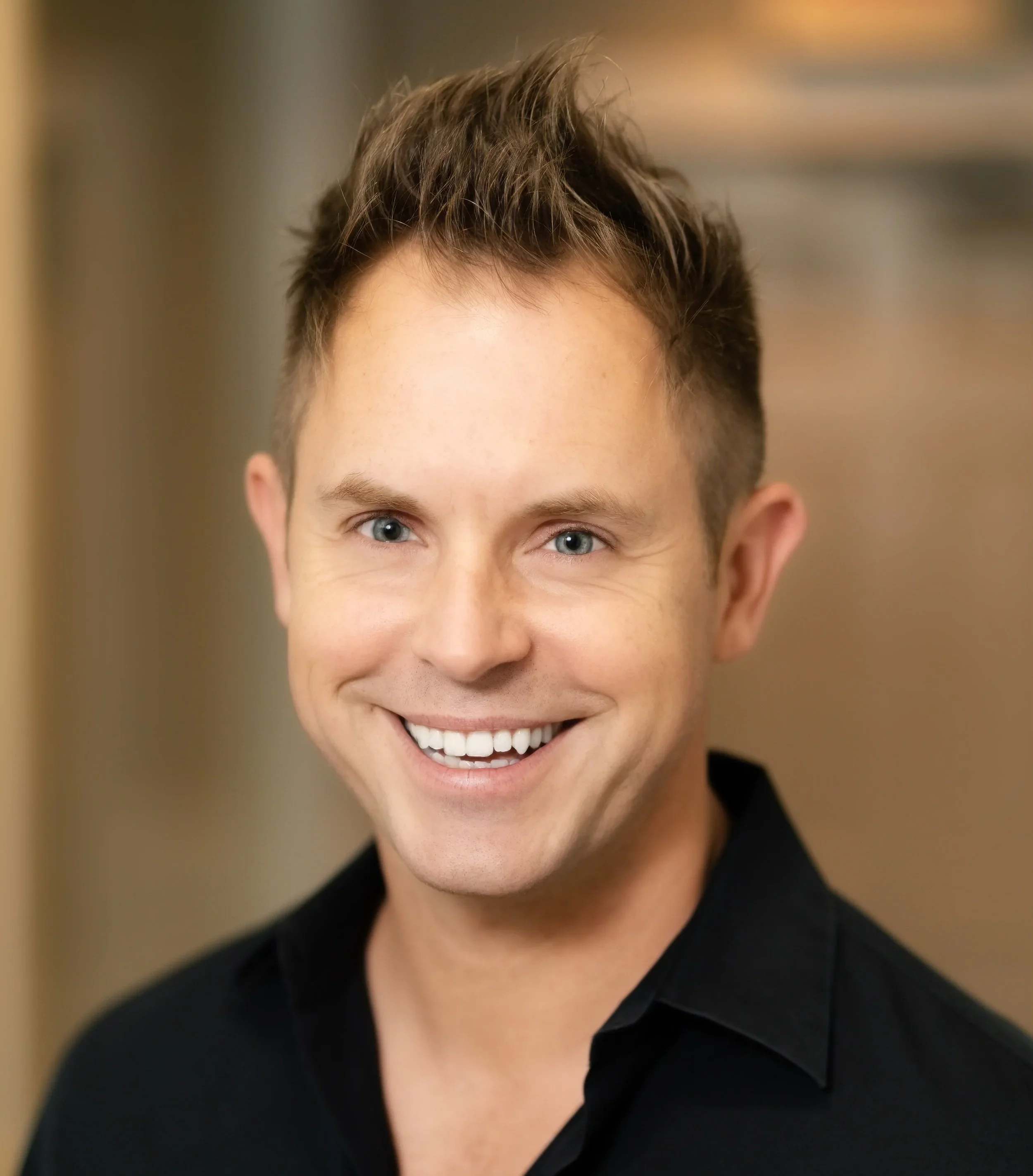 A smiling man with short brown hair and blue eyes, wearing a black shirt, standing indoors against a blurred background.