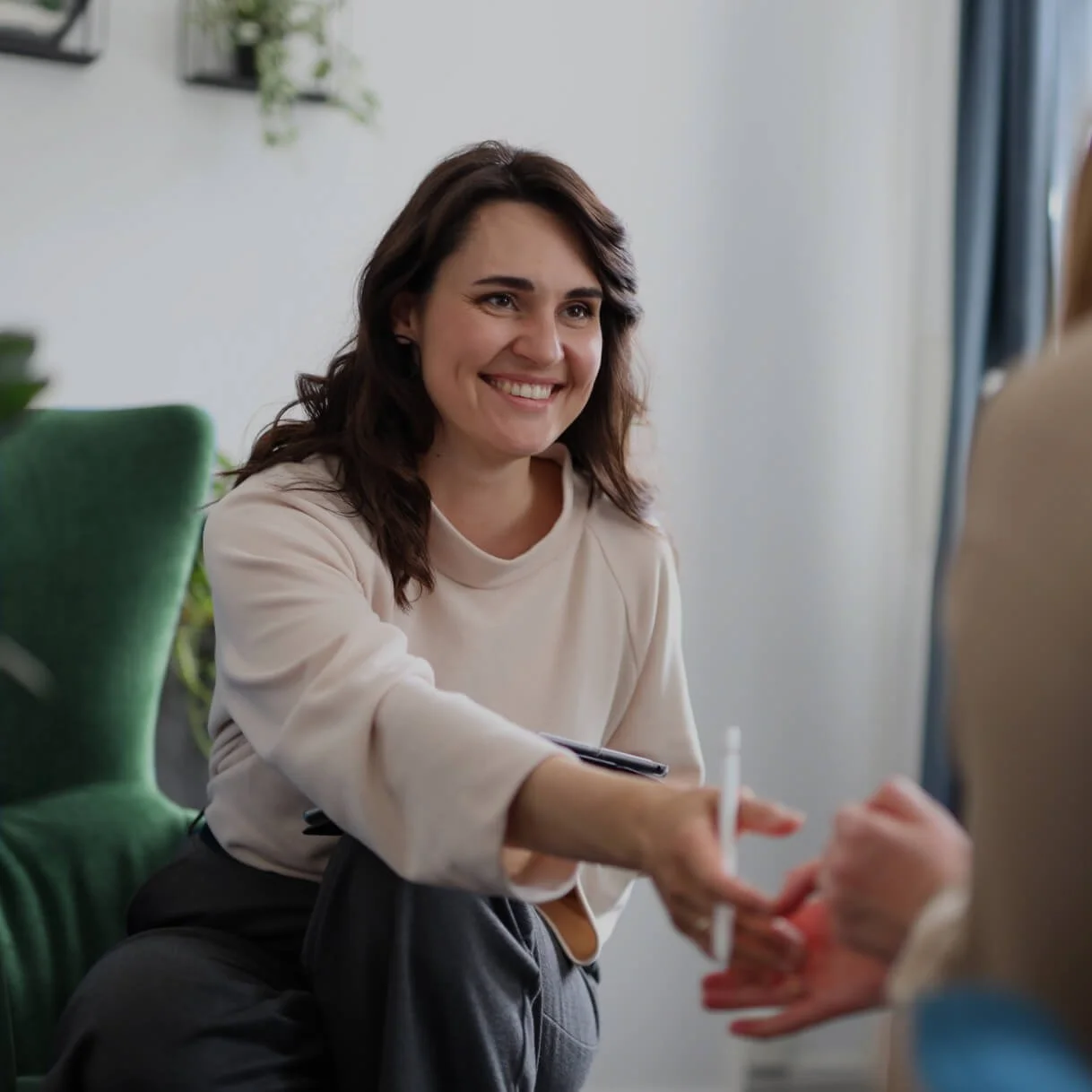 Woman smiling while handing something to another person in a well-lit room with a green chair and a plant in the background.