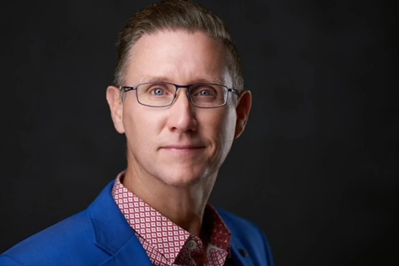 A portrait of a man with glasses, short brown hair, wearing a blue blazer and a red patterned shirt, against a dark background.