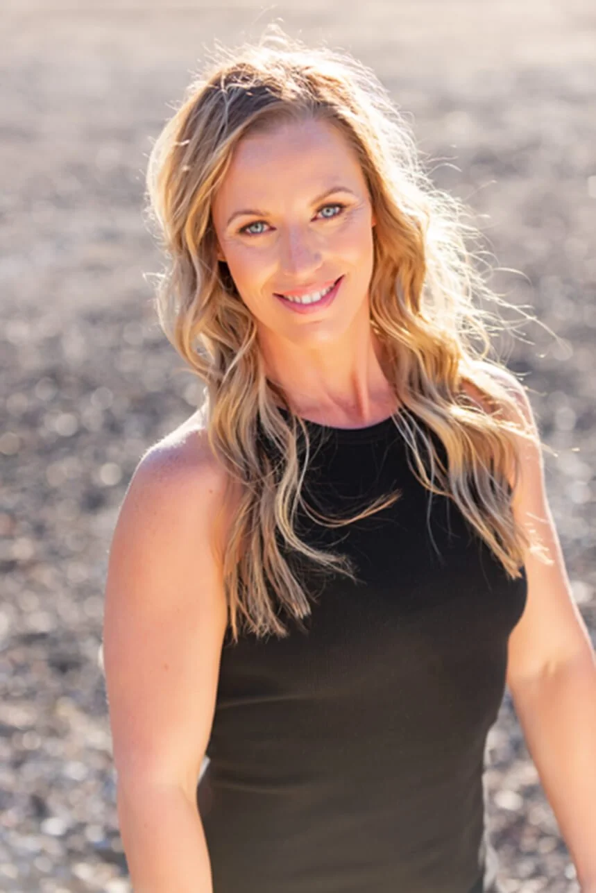 A woman with wavy blonde hair and blue eyes smiling at the camera outdoors in a black sleeveless top with sunlight behind her.