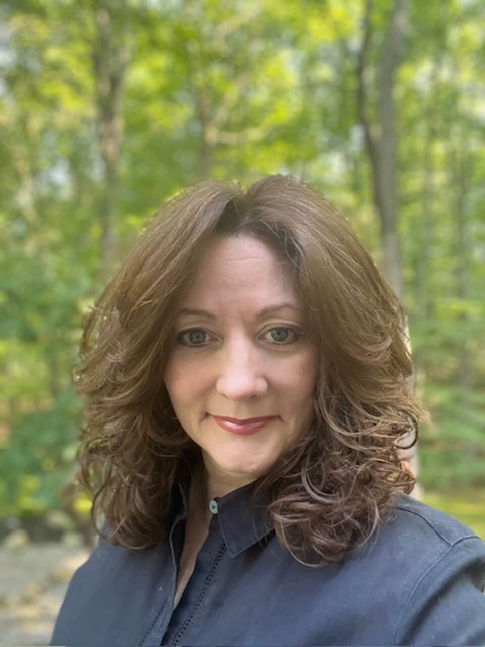 A woman with wavy, shoulder-length brown hair, wearing a dark shirt, smiling outdoors with a green wooded background.