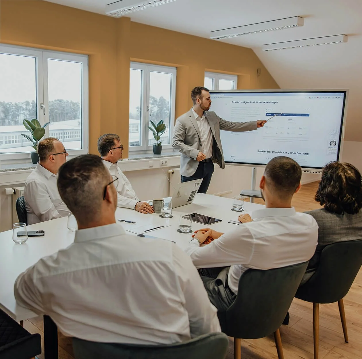 A business consultant presents to a group of professionals seated at a conference table in a bright room with large windows and potted plants, using a large digital screen for the presentation.