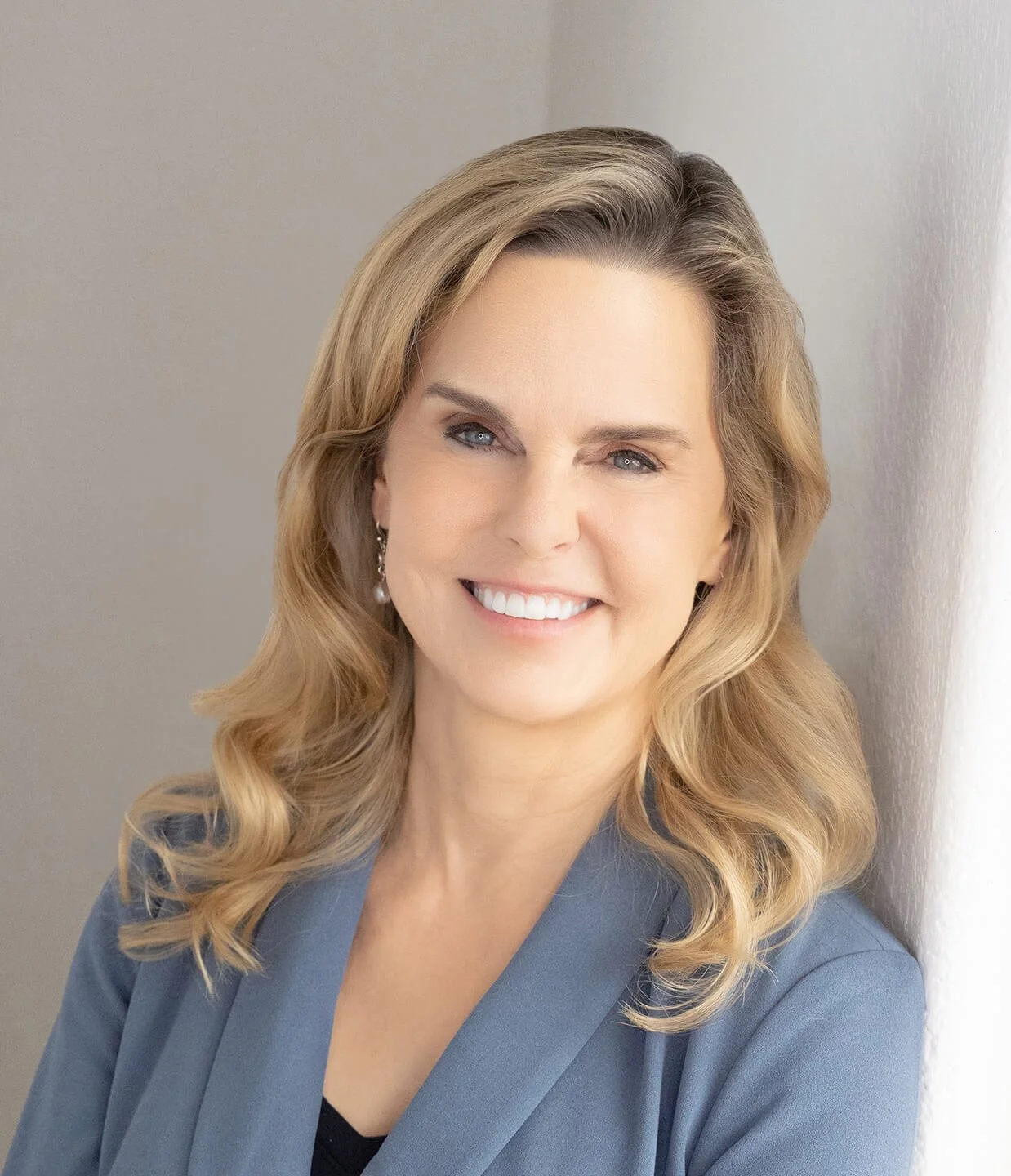 A smiling woman with blonde wavy hair wearing a grey blazer and pearl earrings, leaning against a light-colored wall.
