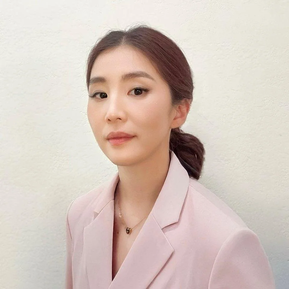 Portrait of a young woman with brown hair in a low ponytail, wearing a light pink blazer and a gold necklace, posing against a plain off-white wall.