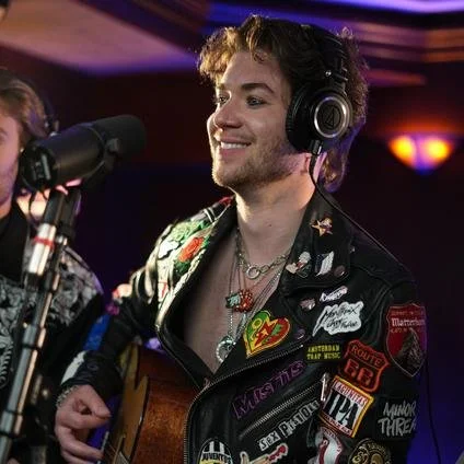 A young man with curly hair singing into a microphone in a recording studio, wearing headphones and a black jacket decorated with various patches, smiling and playing an acoustic guitar.