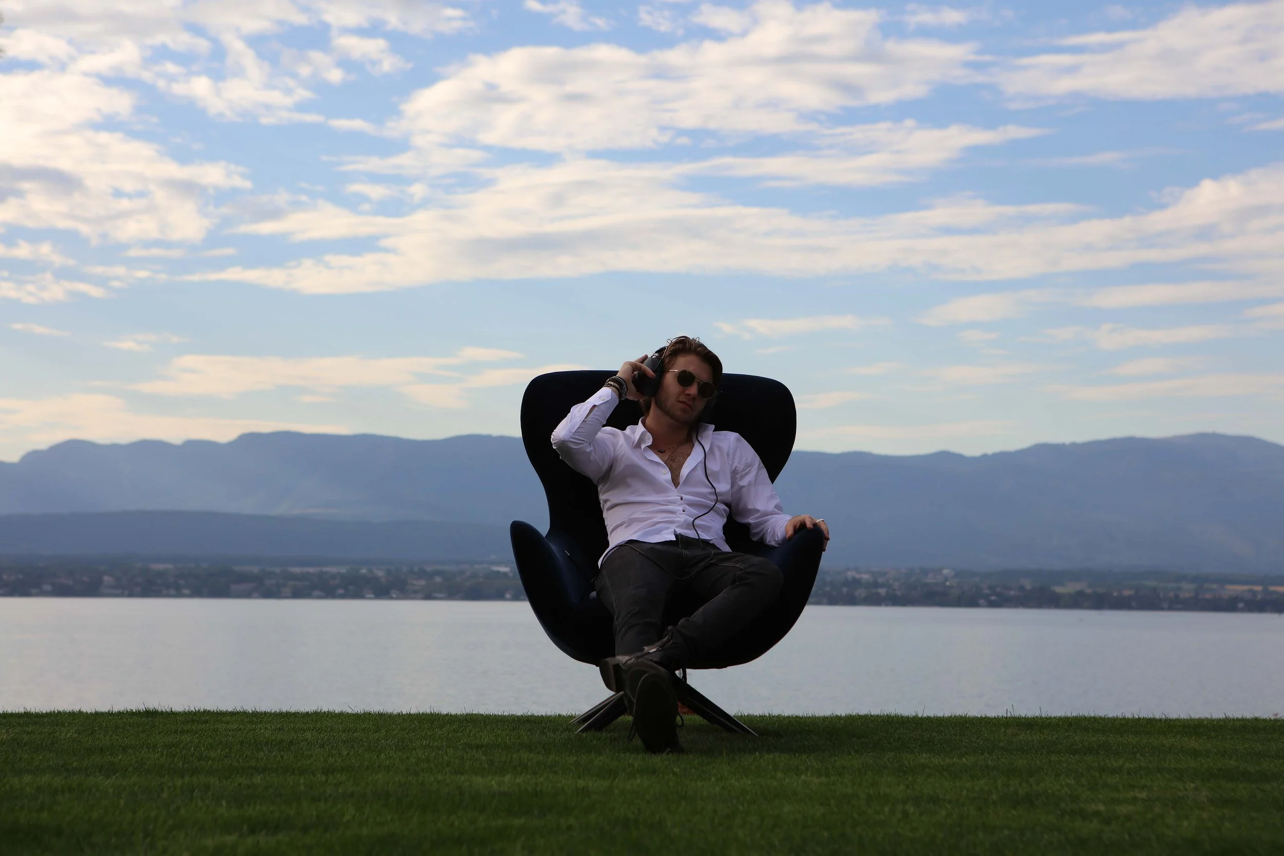 A man with long hair and sunglasses sitting in a black modern chair on a grassy area near a body of water, with mountains in the background and a sky with scattered clouds.