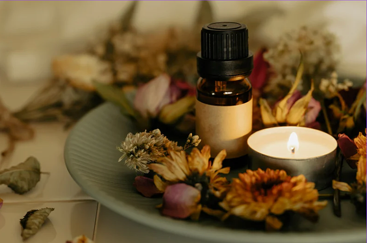 A small amber glass bottle with a black cap, surrounded by dried flowers and a lit tea light candle on a white platter, with additional dried flowers and petals in the background.