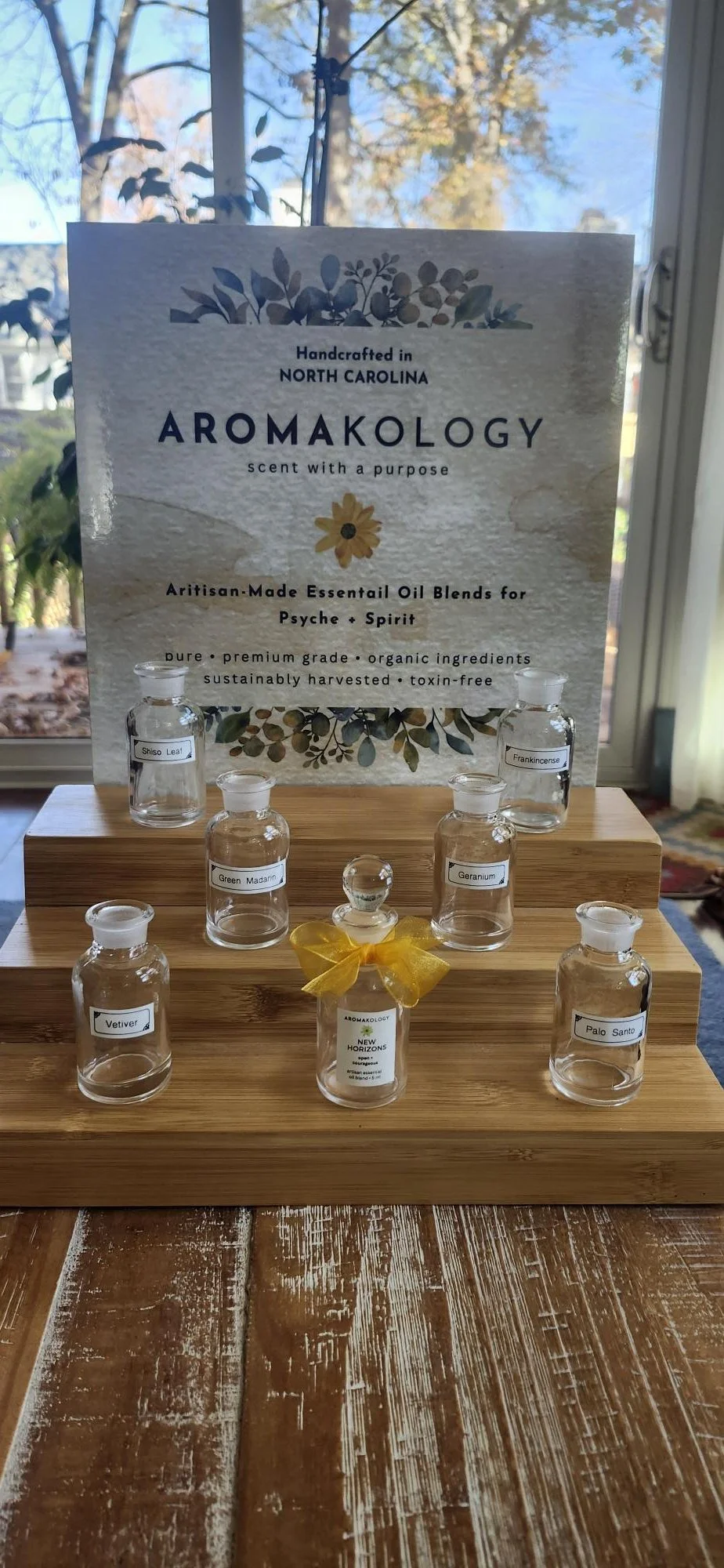 Display of seven glass bottles with labels of essential oils, arranged on a wooden tray in front of a sign that reads "Aromakology, handcrafted in North Carolina, scent with a purpose." The bottles are labeled Shiso Leaf, Frankincense, Green Mandar, Geranium, Vetiver, Palo Santo, and a bottle with a yellow ribbon labeled New Horizons, with a background of trees and a window.