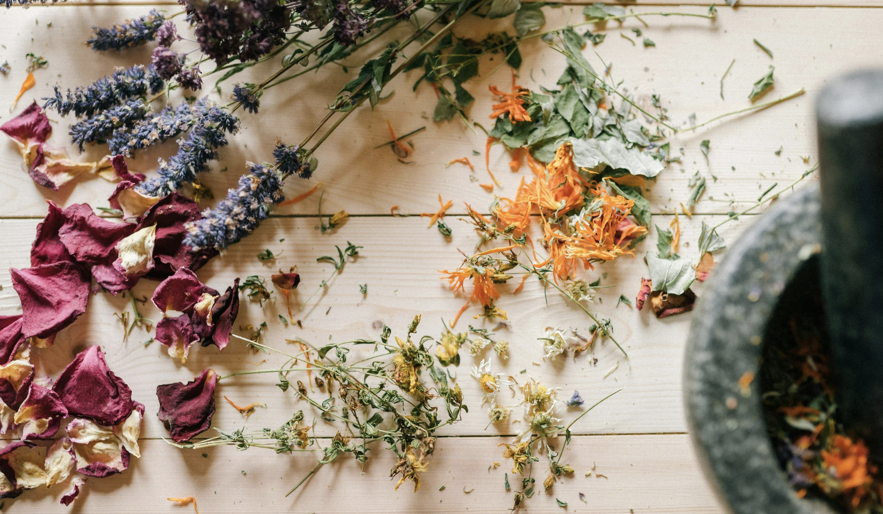 Dried flowers and herbs on a wooden surface with a stone mortar and pestle containing more dried flowers and herbs on the right.