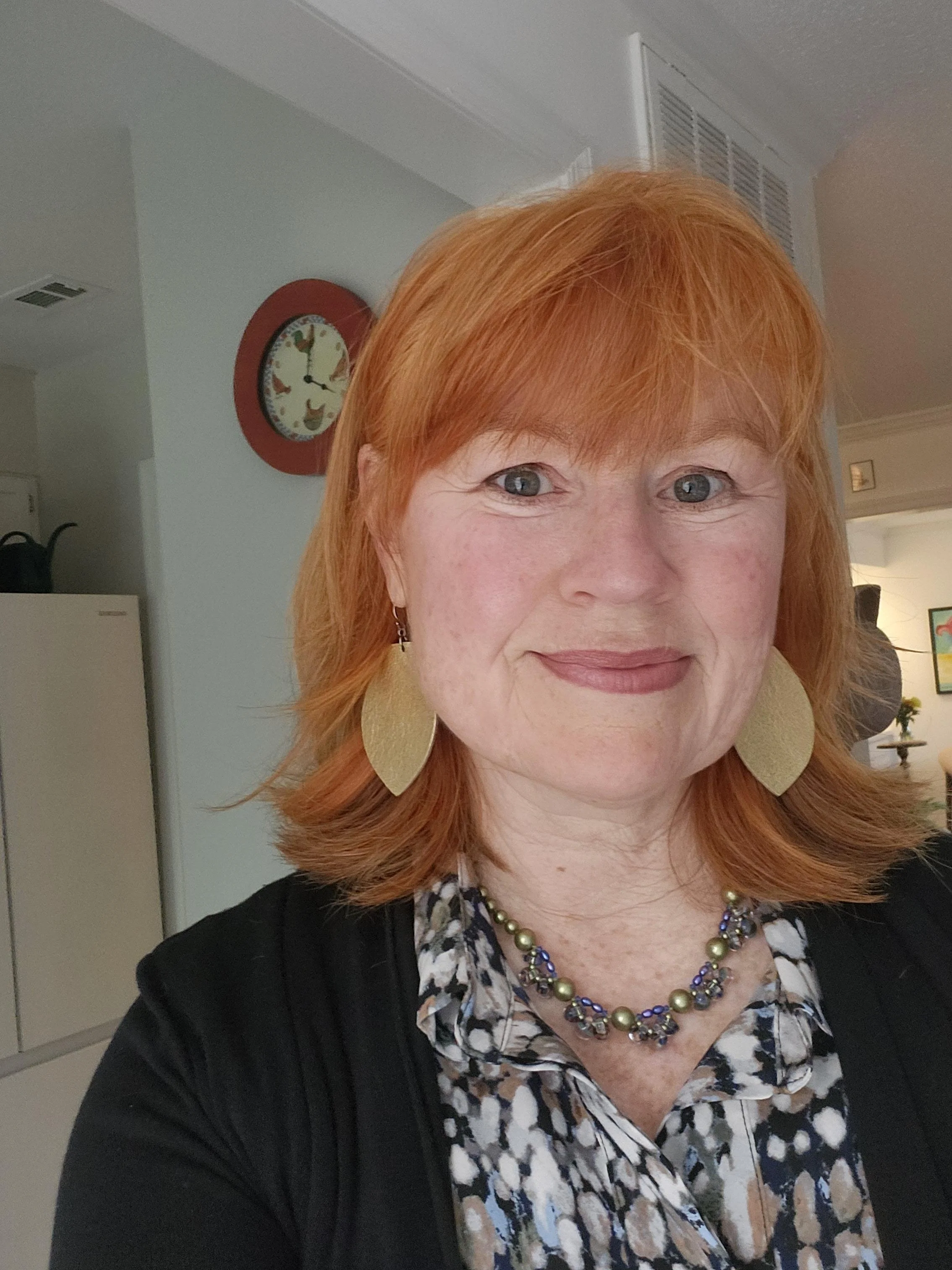 Close-up of a woman with red hair, wearing earrings and a necklace, smiling slightly, indoors with a kitchen and colorful wall art in the background.