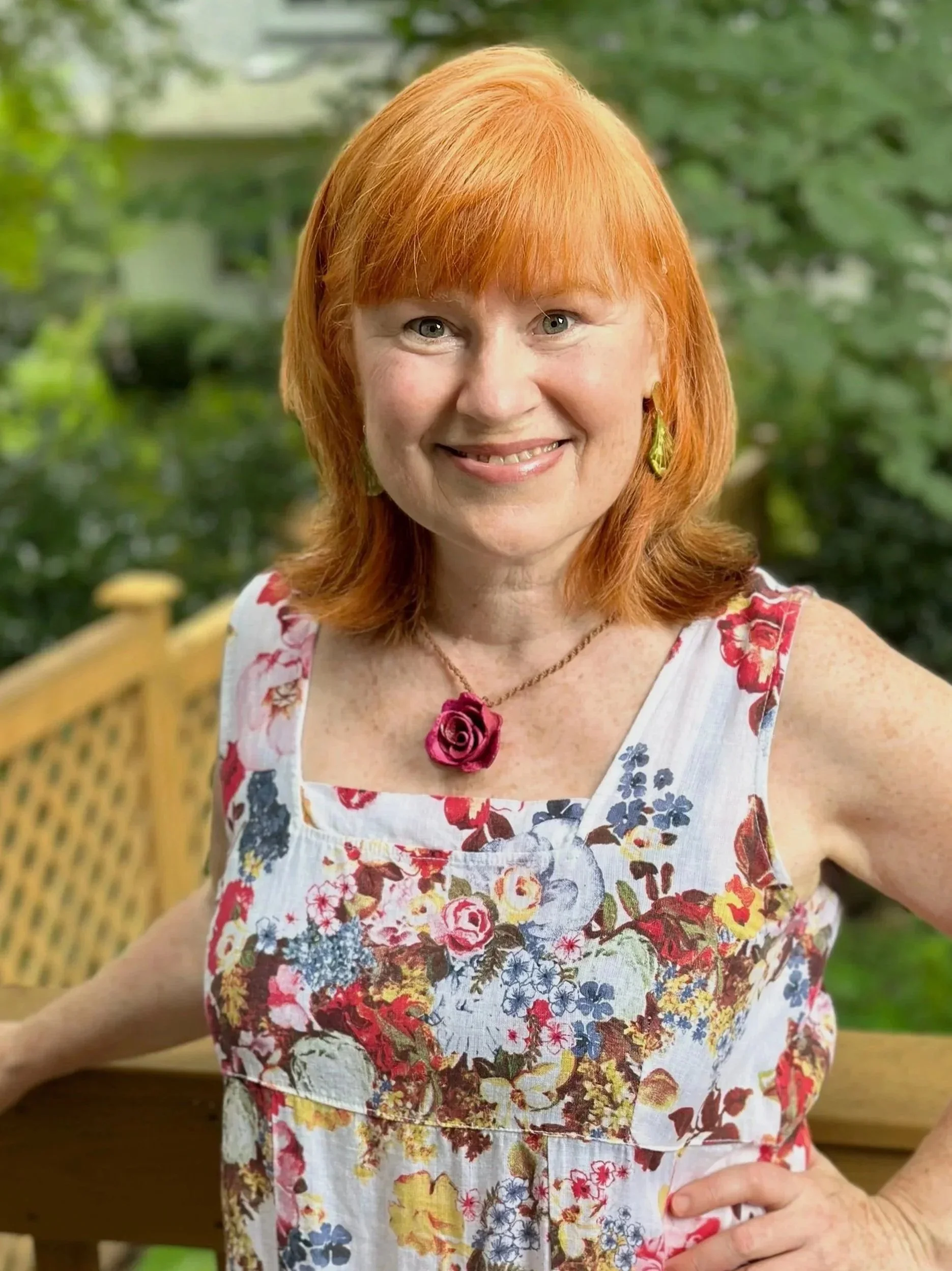 A smiling woman with red hair wearing a sleeveless floral dress, a pink flower necklace, and gold earrings, standing outdoors on a wooden deck with green trees in the background.
