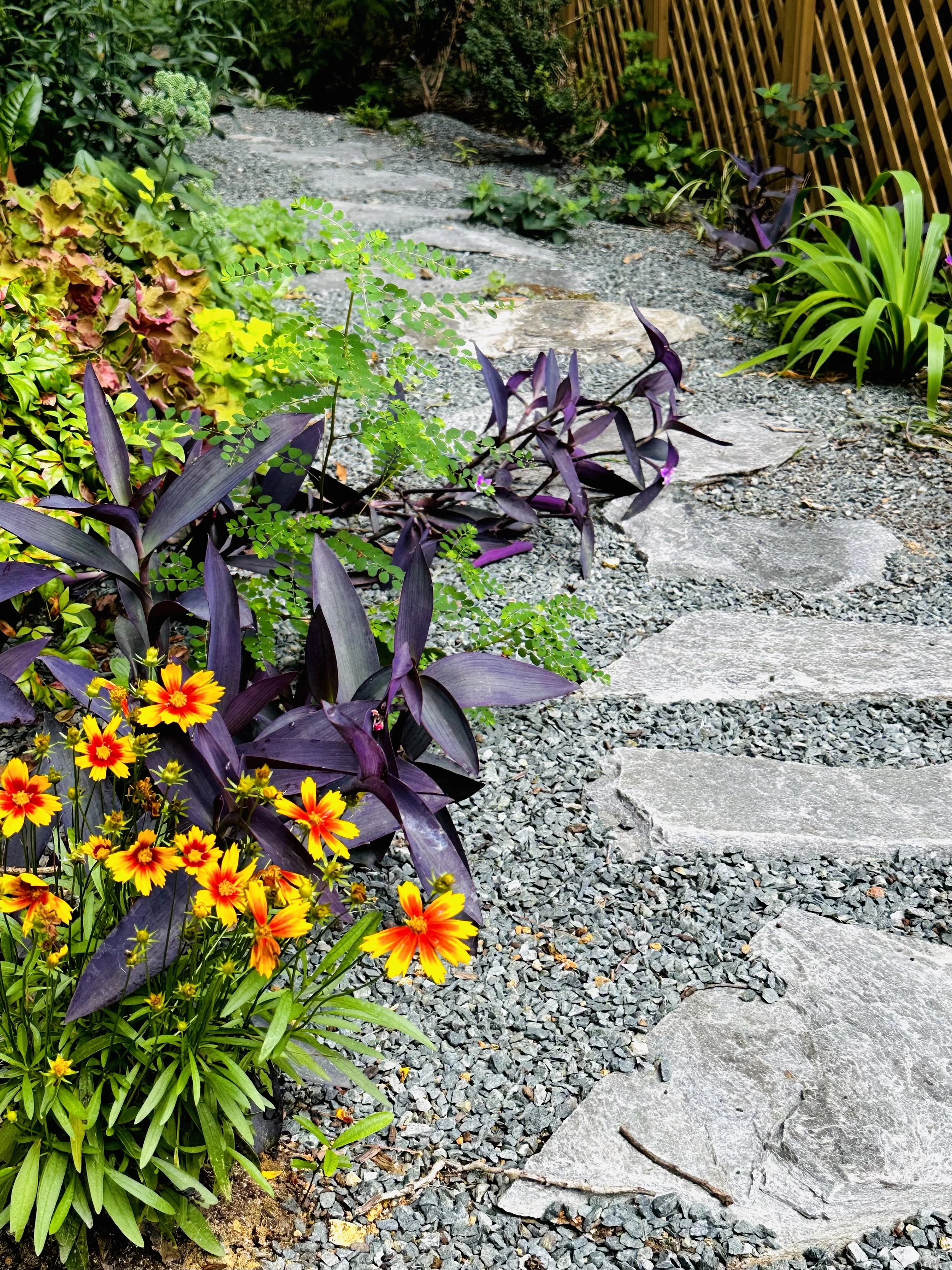 A garden pathway made of stepping stones and gravel, bordered by colorful flowers and green foliage, with a wooden lattice fence on the right side.